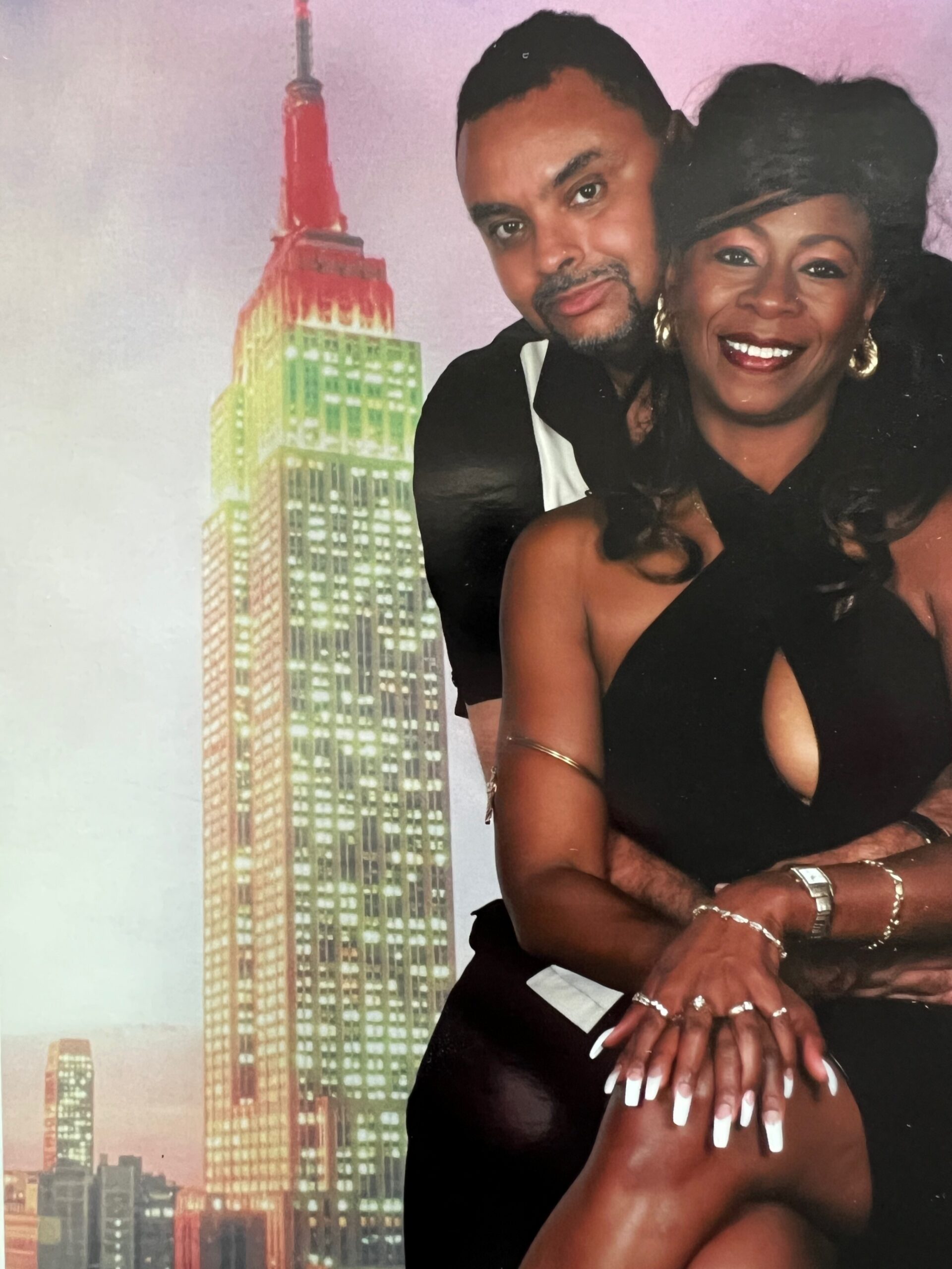 A smiling couple posing close together in front of a city backdrop, the woman wearing a black dress and gold jewelry.