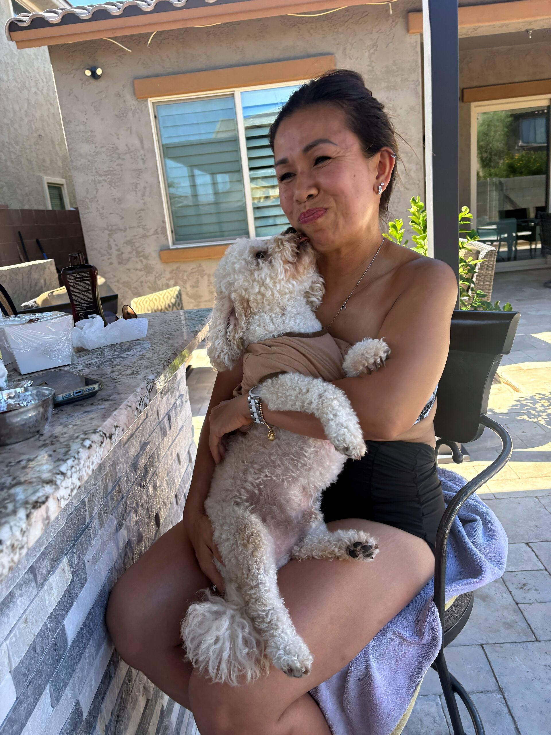 Smiling woman in a strapless top cuddling a small curly white dog on a backyard patio, with a counter and outdoor furniture nearby.