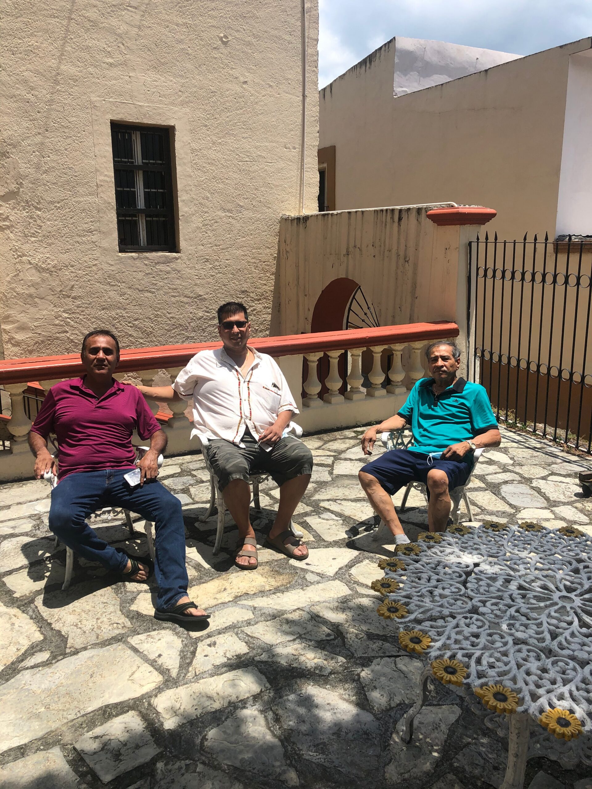 Three men sit on white patio chairs on a sunlit stone terrace beside beige walls.
