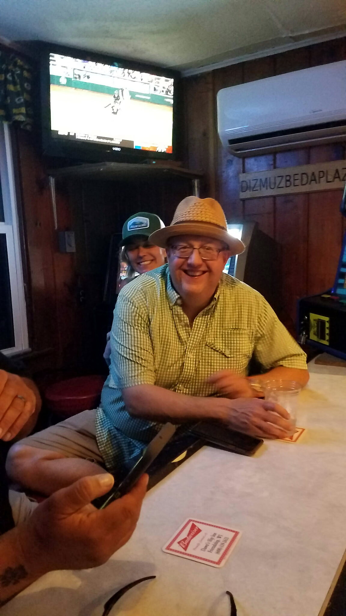 Smiling man in a straw hat and glasses sits at a table in a cozy bar, with a TV on the wall behind and another person in a cap smiling beside him.