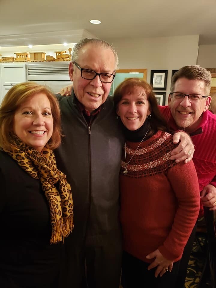 Four adults posing together indoors, smiling with arms around each other in a warm home setting.