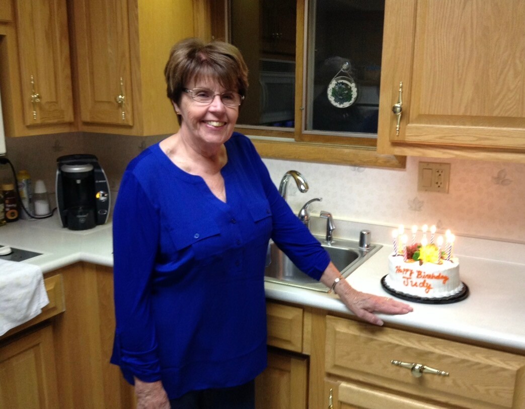 Smiling woman in a blue blouse stands at a kitchen counter next to a birthday cake with lit candles.