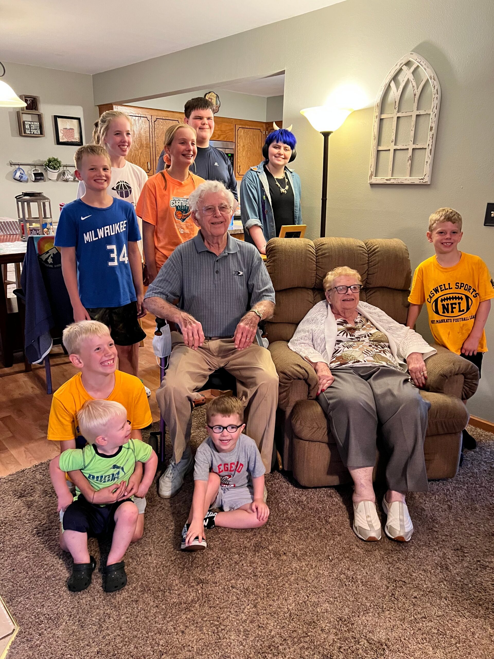 Group of children posing with an elderly man in a wheelchair and an elderly woman in a recliner in a living room; everyone smiling, kitchen cabinets visible in the background.