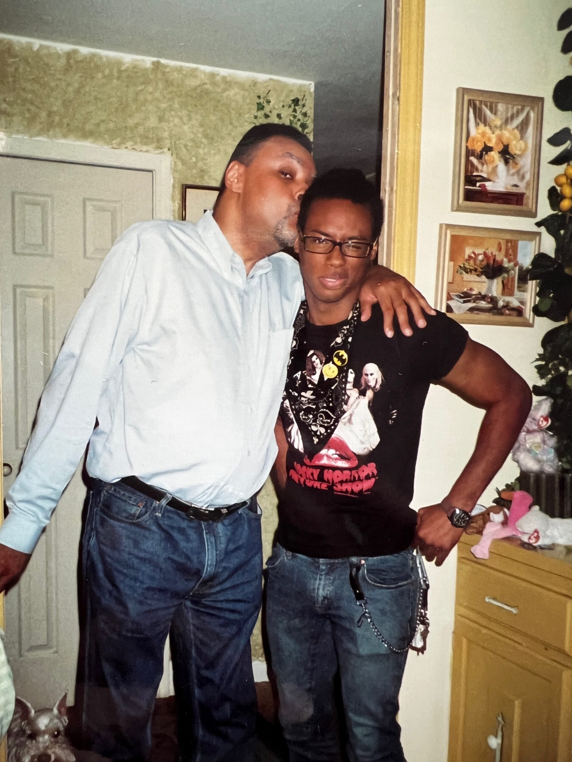 Two men posing in a home, one kissing the other on the cheek with an arm around his shoulders.
