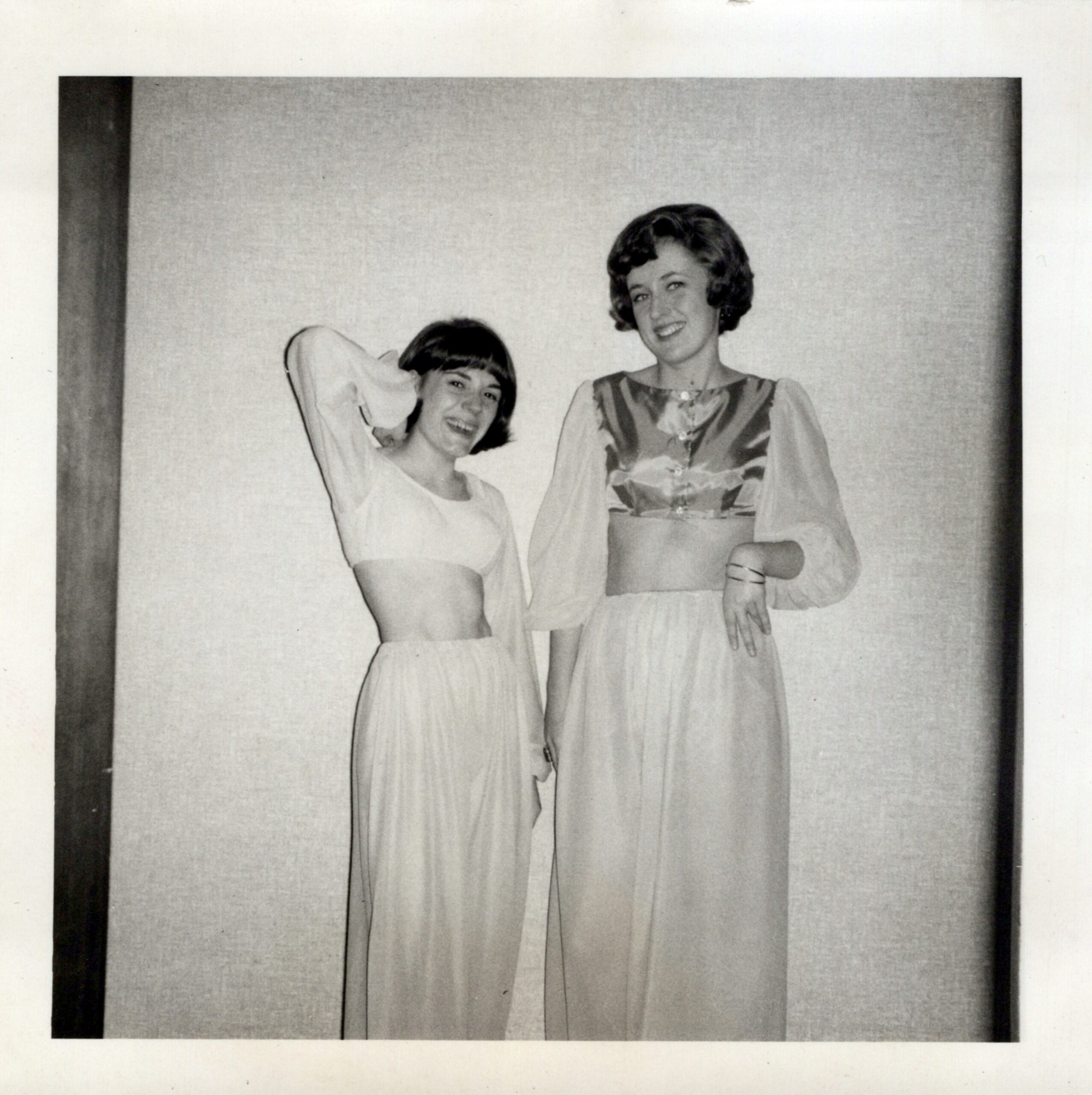Two women in vintage dresses pose and smile for the camera, standing side by side in a simple backdrop.