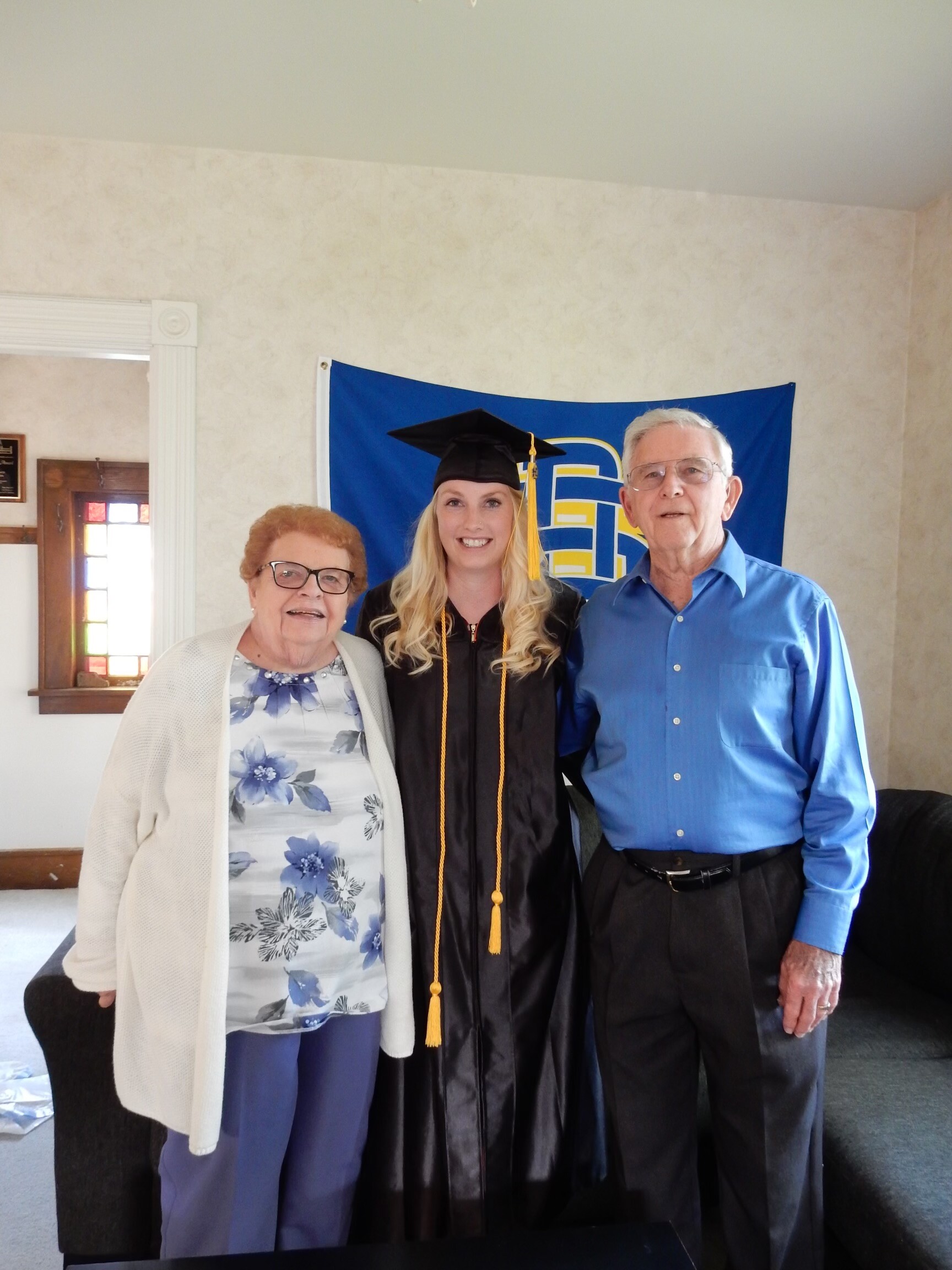 Graduate in cap and gown with gold tassel stands between an elderly woman and man indoors, blue banner in background.