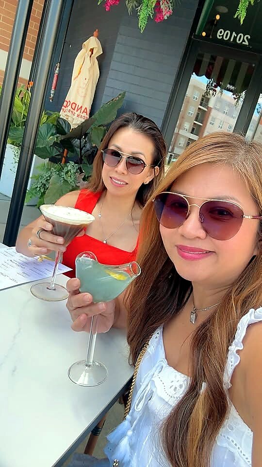 Two women at an outdoor cafe, wearing sunglasses and holding cocktails; one in a red top with a foamy drink, the other in a white blouse with a blue-green cocktail.