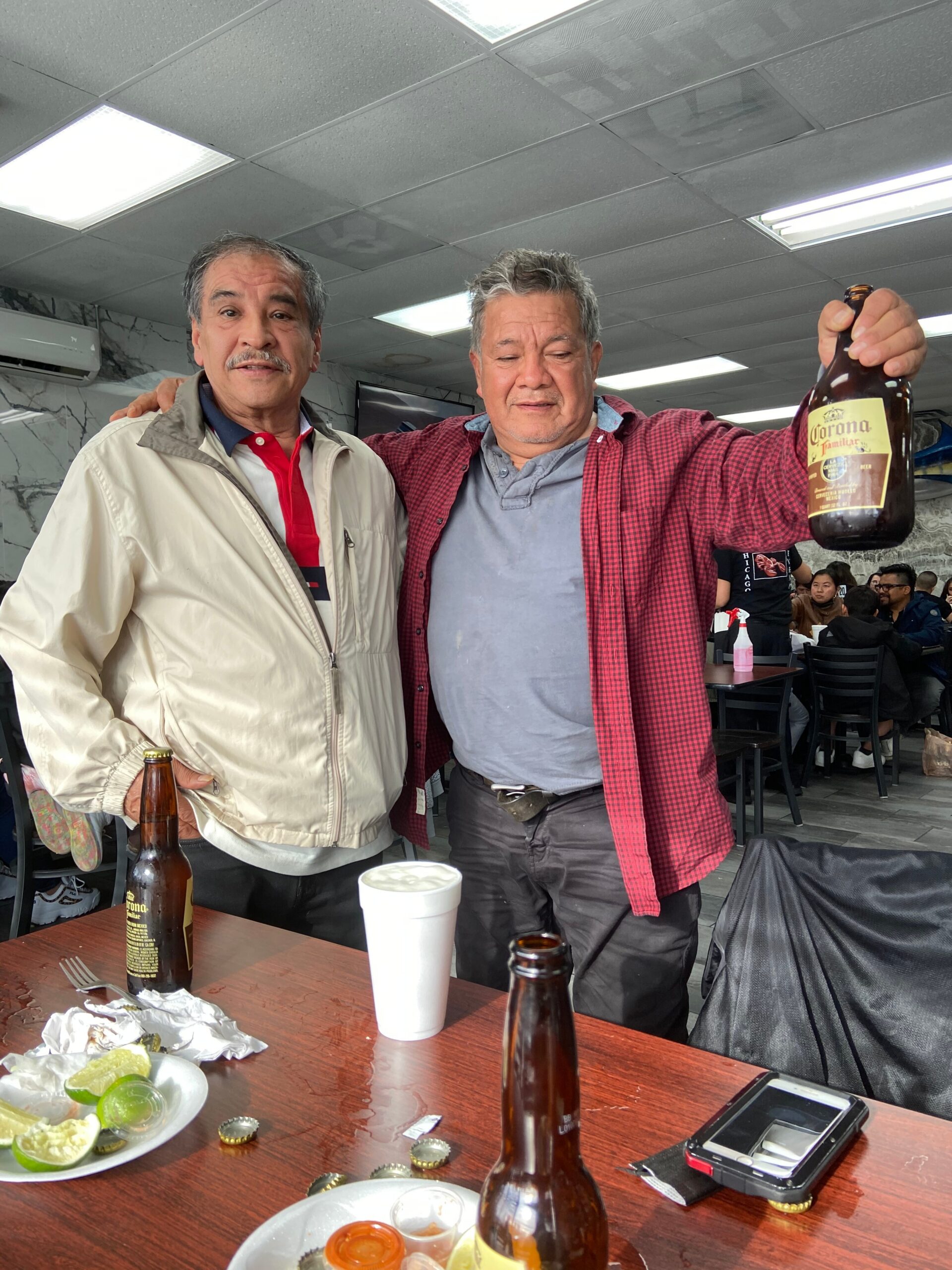 Two men posing together in a casual restaurant; one raises a Corona beer bottle toward the camera.