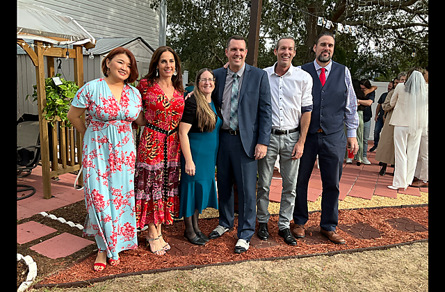 Six adults pose together outdoors at a celebration, smiling for the camera; three women on the left in colorful dresses and three men on the right in semi-formal attire, against a garden venue backdrop.