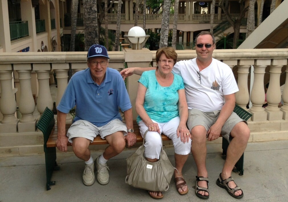 Three adults sit on a bench outdoors, smiling at the camera with a stone balustrade and palm trees behind them.