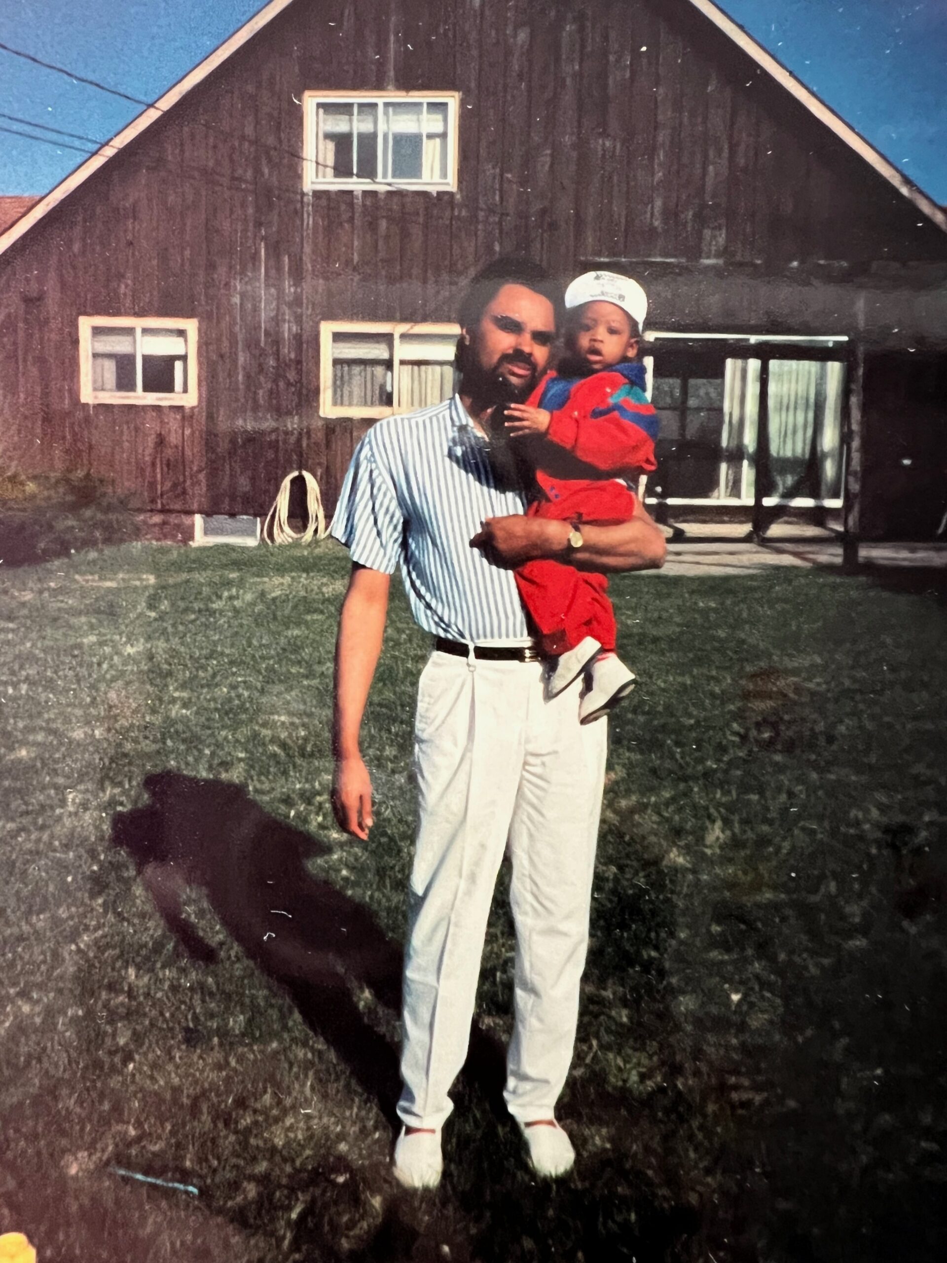 Man with a beard in a striped shirt holds a toddler in red clothing outside a dark wooden house.
