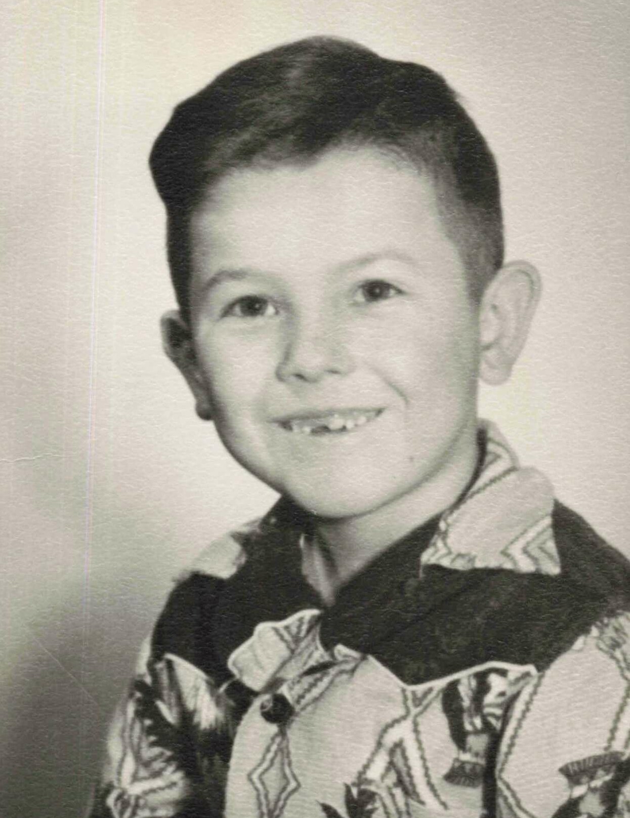 Black-and-white photo of a smiling boy with short dark hair wearing a patterned button-down shirt, facing the camera.