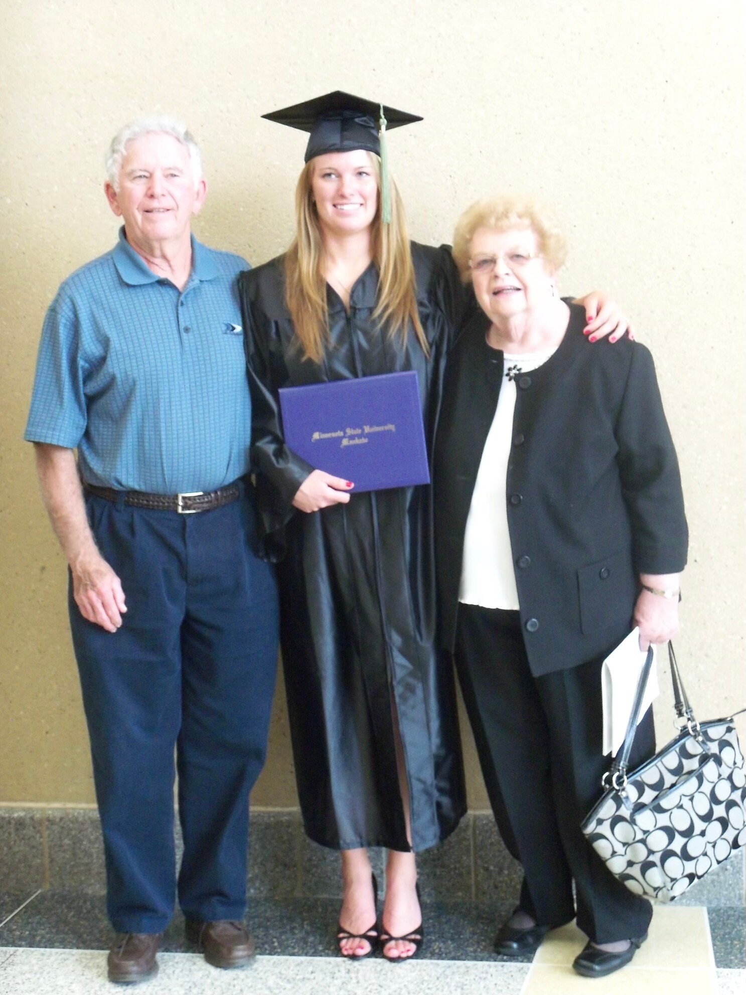 Graduate in cap and gown holding a purple diploma folder, posing with an elderly man and woman in a hallway.