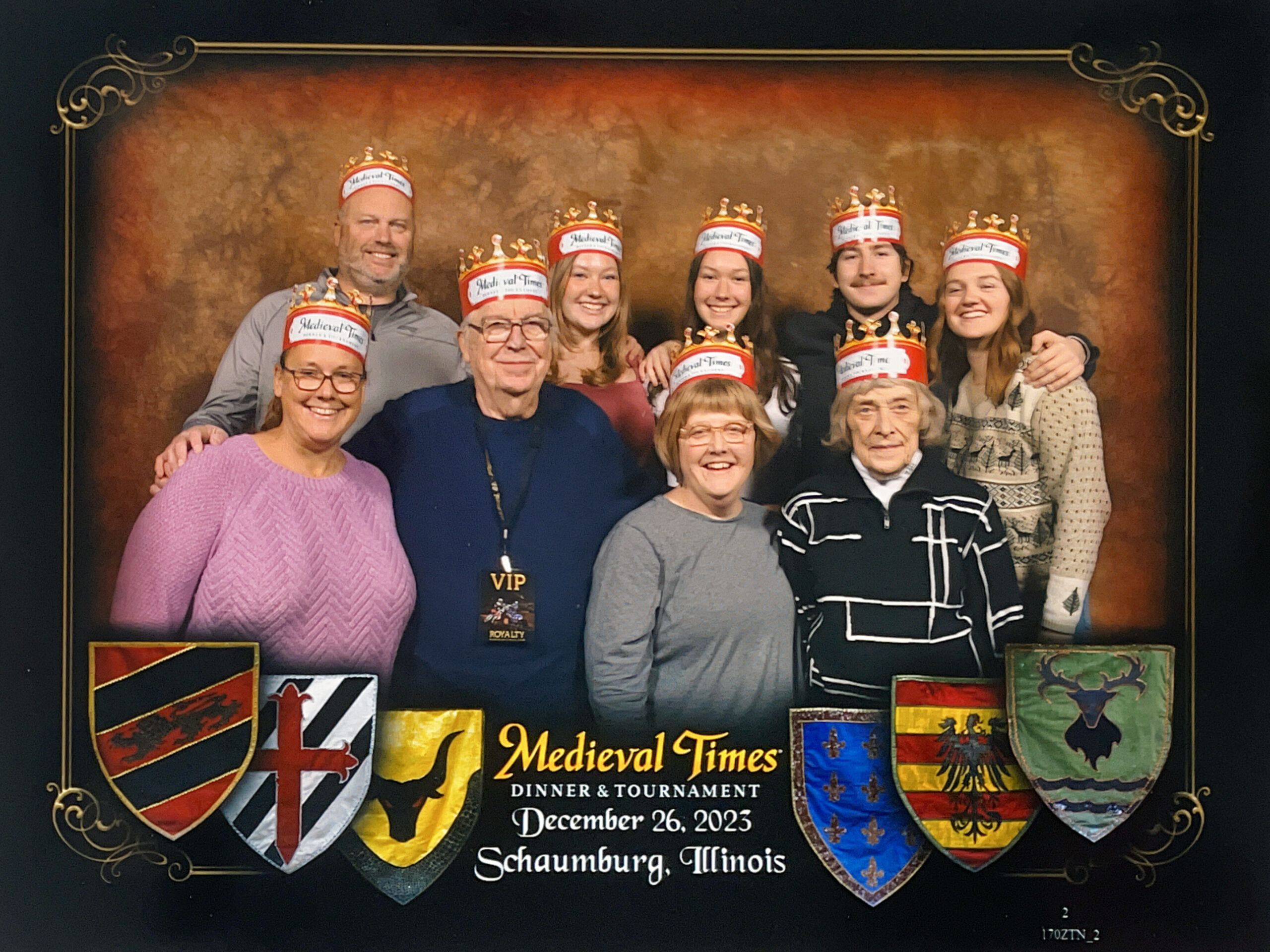 Group of people wearing paper crowns posing in a medieval-themed display with coats of arms and a banner bannered 'Medieval Times'