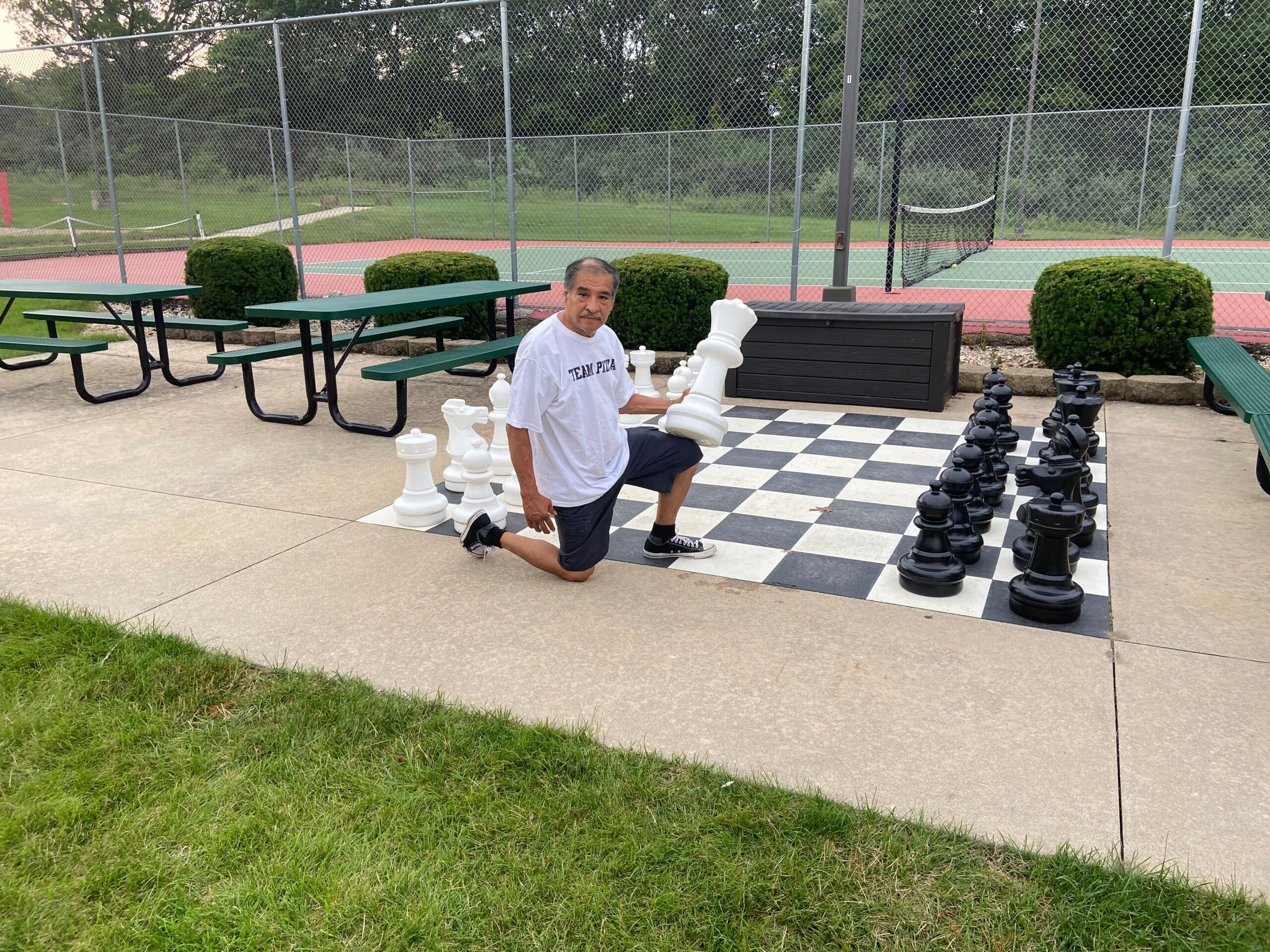 Man kneeling on a huge outdoor chessboard, examining a white king among oversized chess pieces.