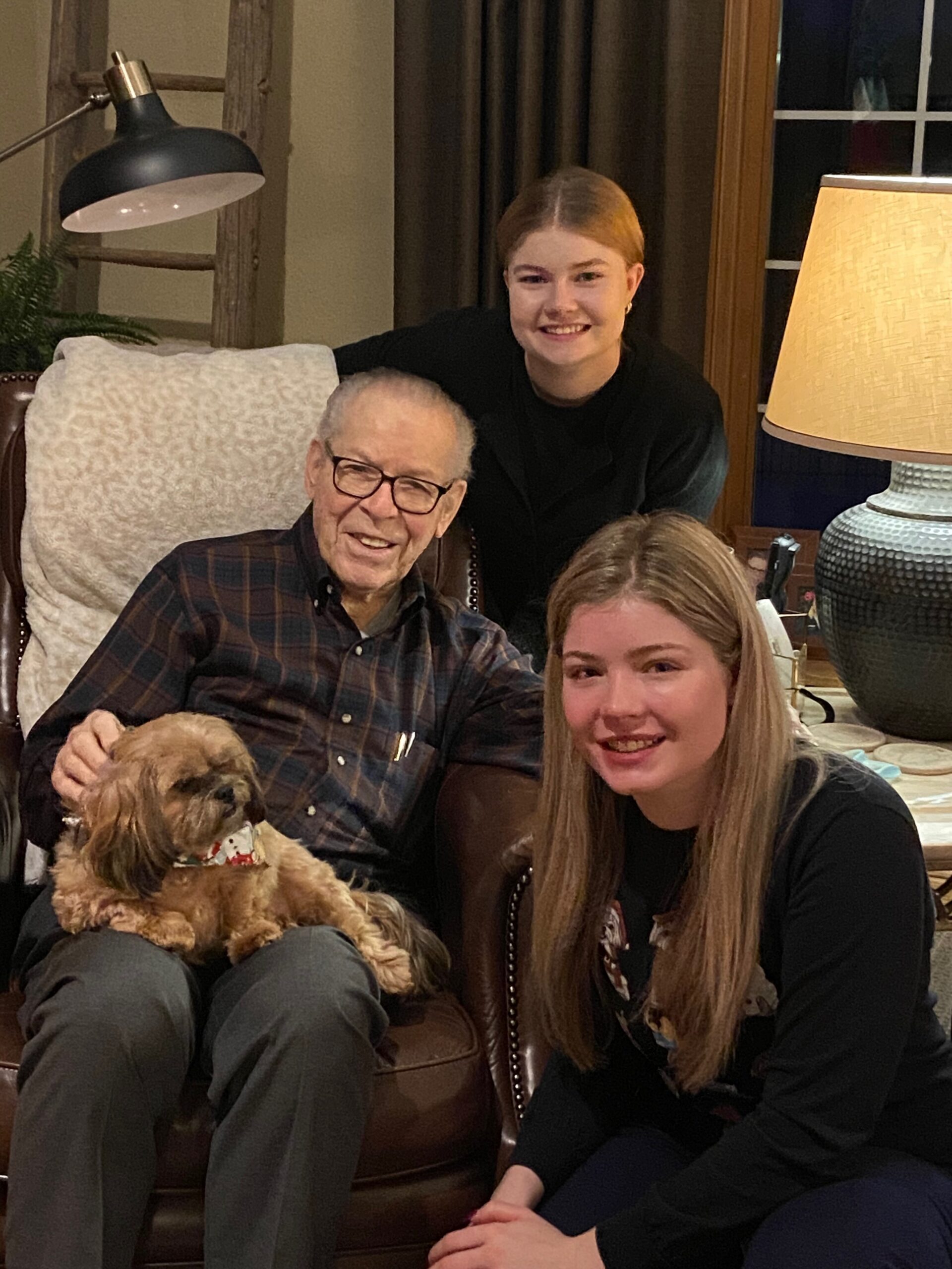 Smiling elderly man with glasses in a plaid shirt sits on a leather chair, flanked by two young women and a small brown dog in a cozy living room.