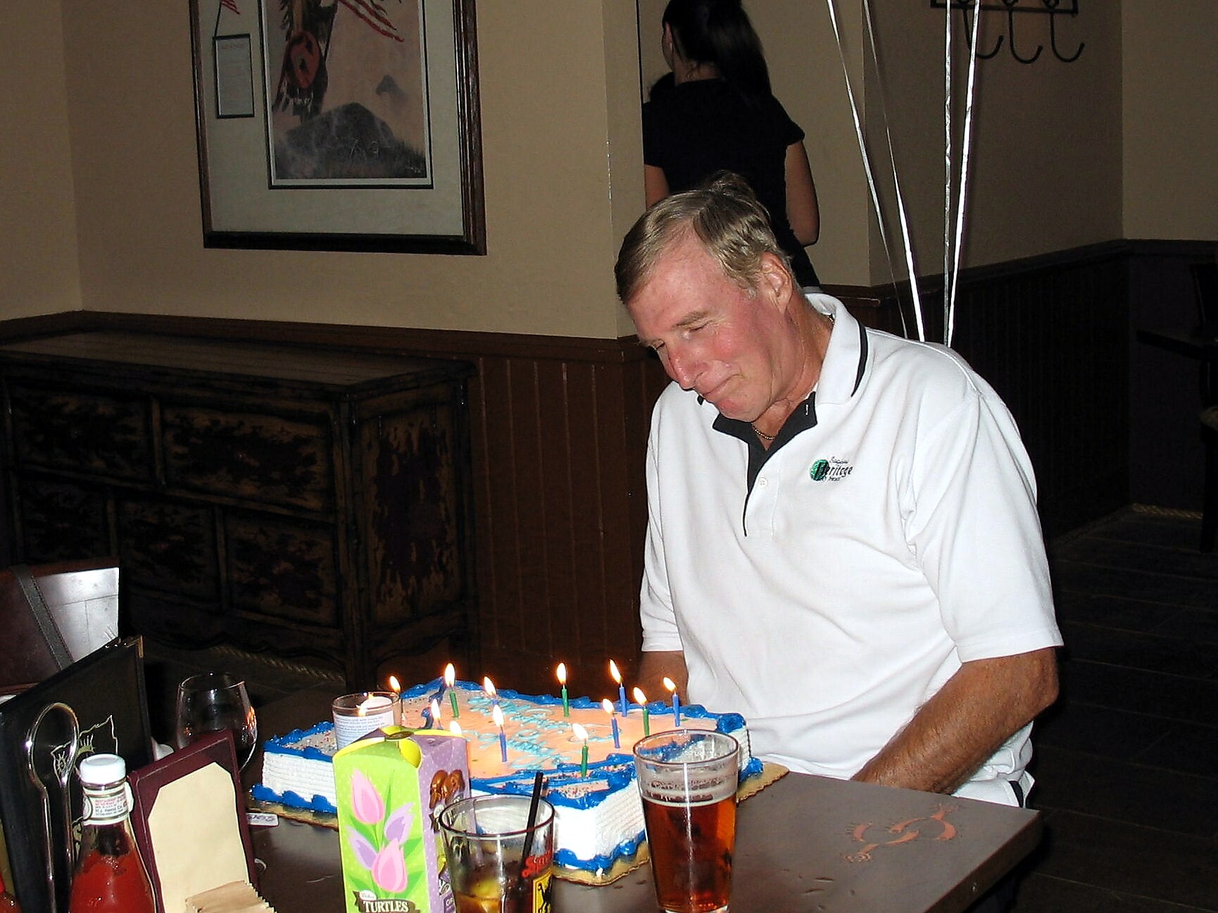 Man in a white polo shirt sits at a table with a blue birthday cake and lit candles in front of him at a restaurant party.