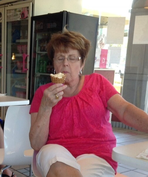 Older woman in a pink blouse sits at a dining area, eating an ice cream cone with eyes closed.