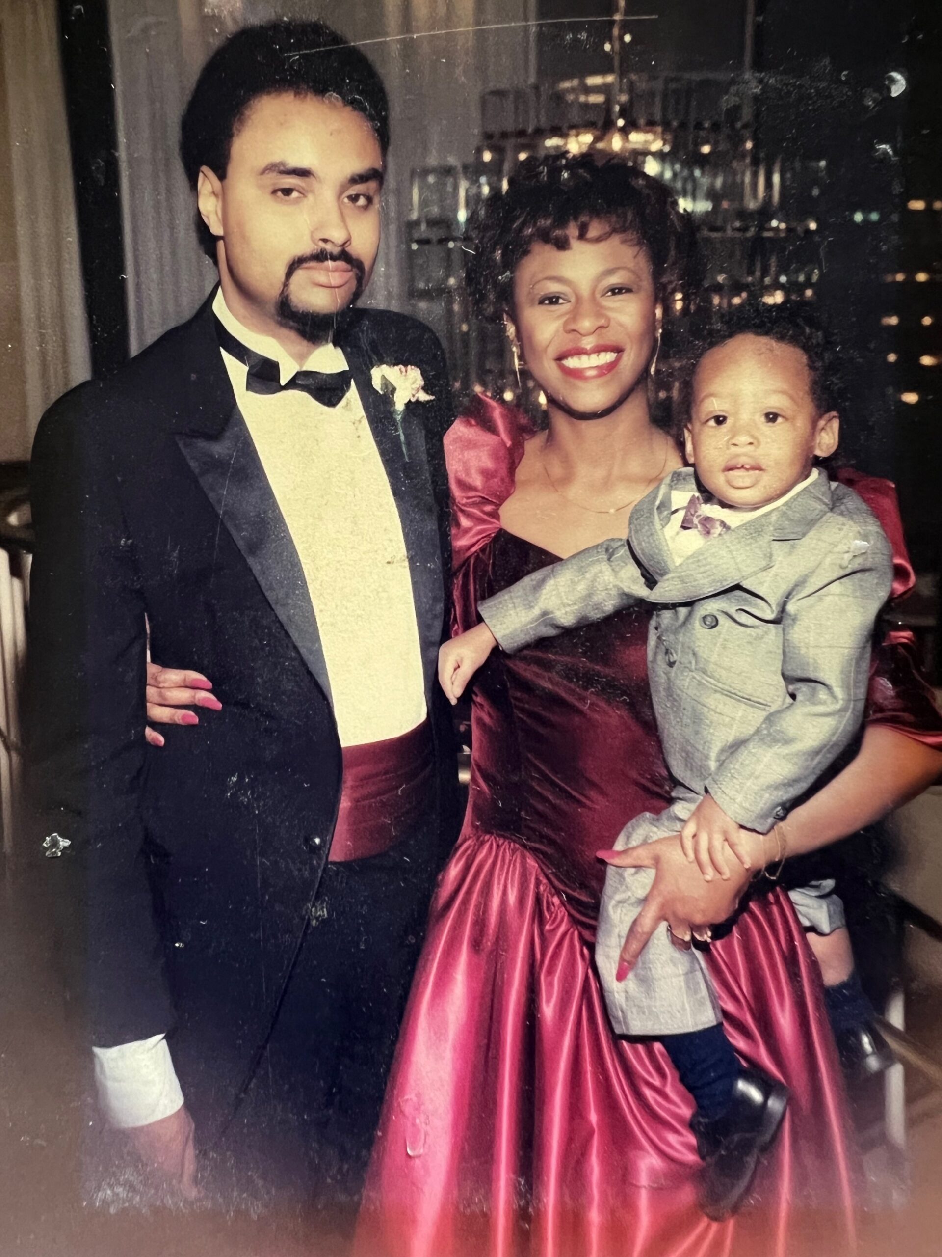 Man in a black tuxedo and bow tie stands beside a smiling woman in a red formal dress, holding a baby in a gray suit underground lighting look.