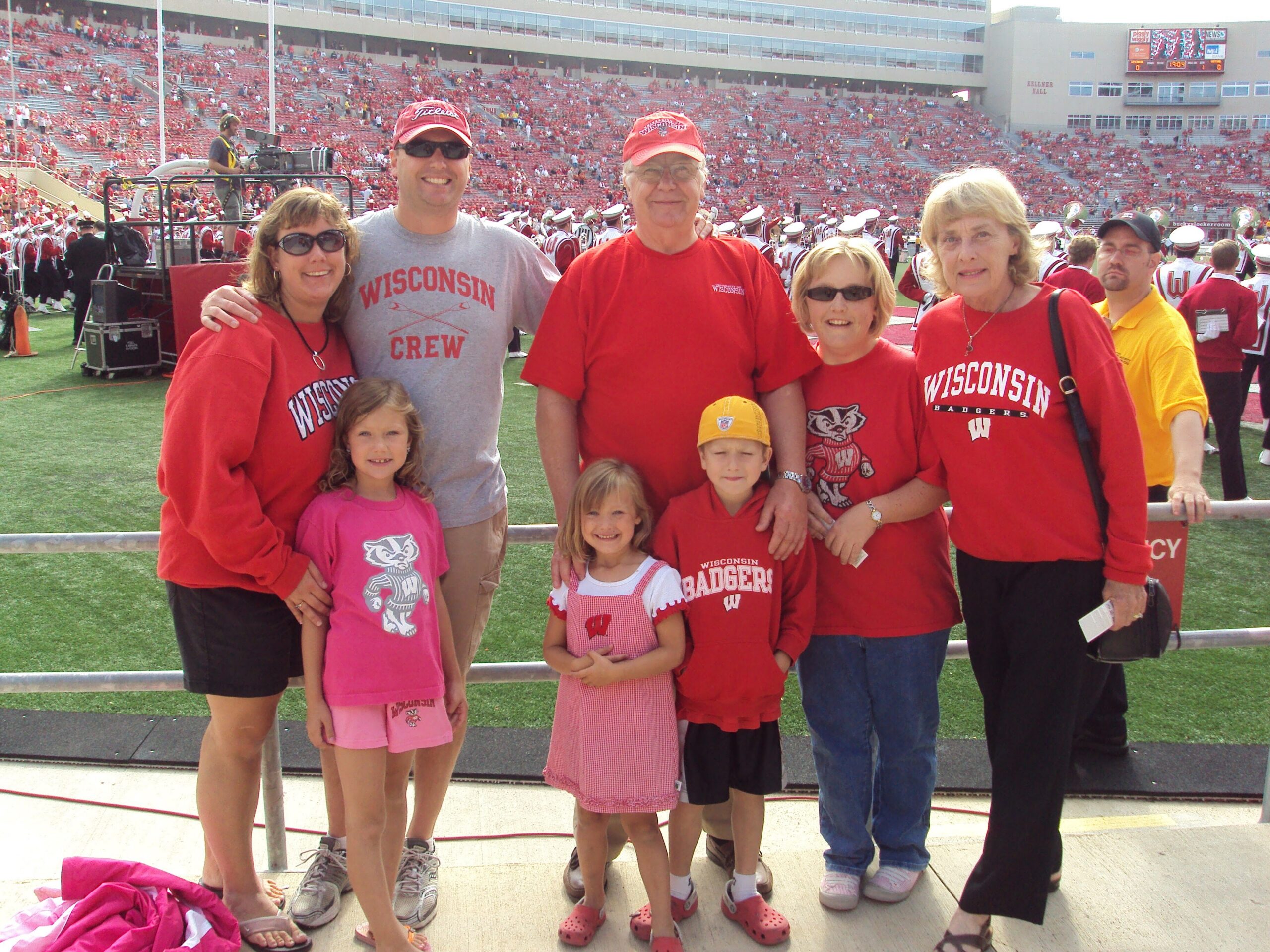 Group of Wisconsin Badgers fans posing on a football field in red shirts, with a large red-packed stadium in the background.
