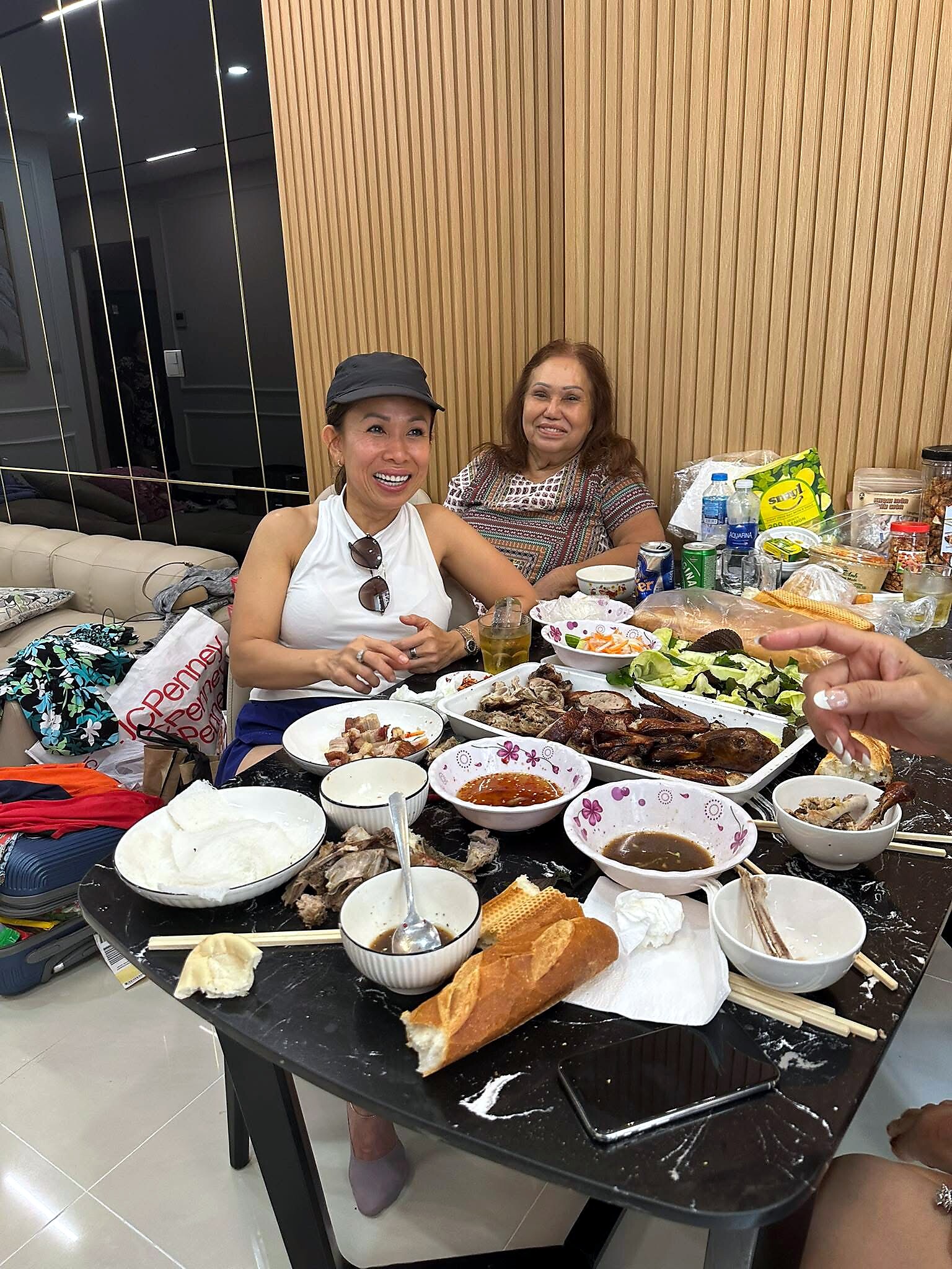 Two women smiling at a dinner party seated around a table filled with roast meat, sauces, bread, and bowls during a casual gathering indoors.