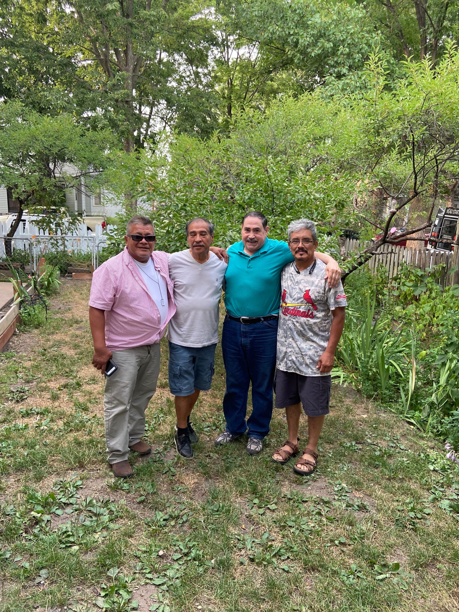 Four men pose together in a backyard garden with trees and shrubs behind them, smiling at the camera.