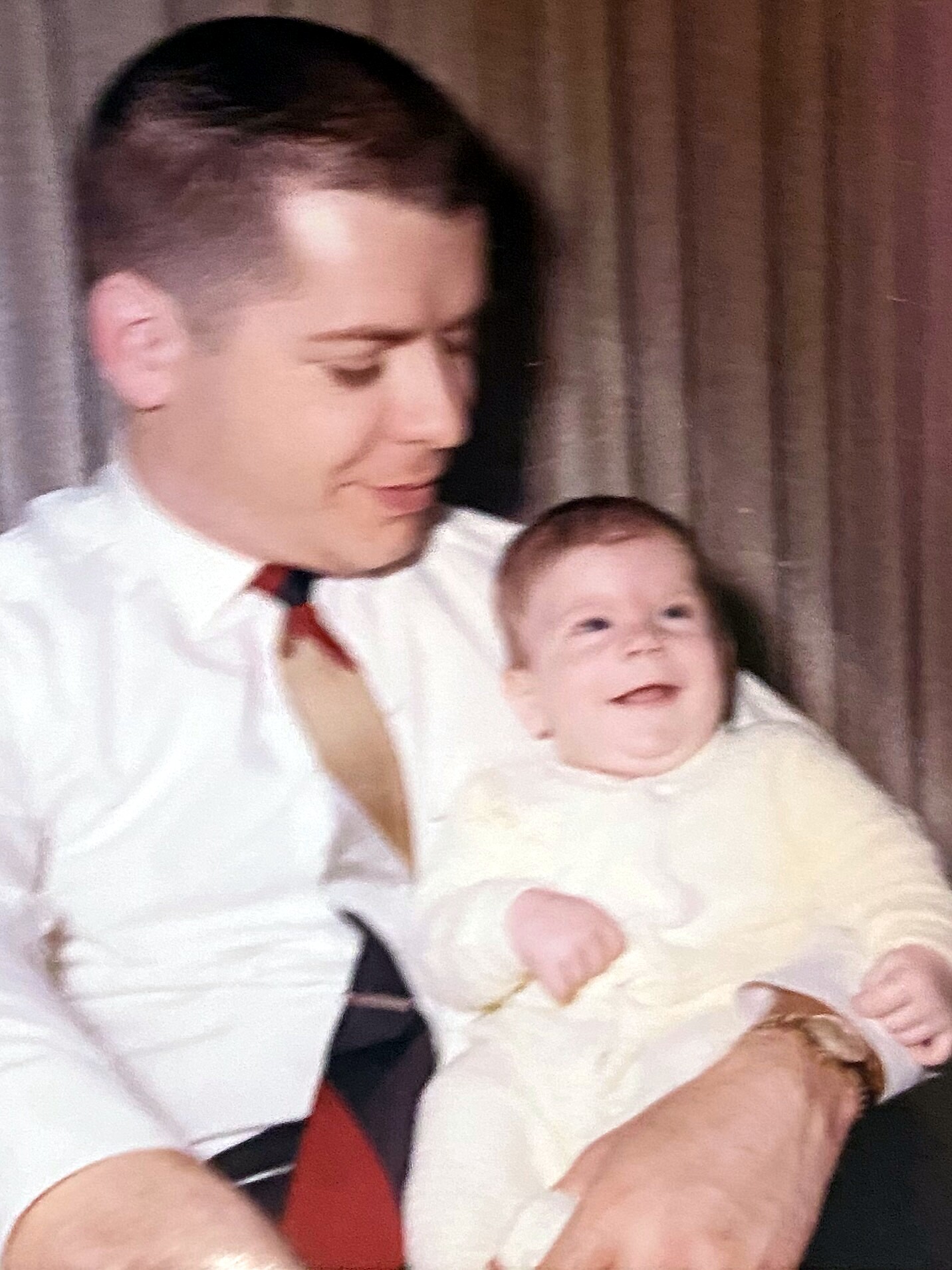 Man in a white shirt and tie holding a smiling baby indoors in front of brown curtains, looking down at the child.