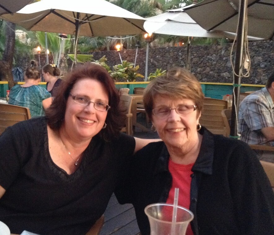Two smiling women sit close together at an outdoor patio with umbrellas, arms around each other, enjoying a meal.
