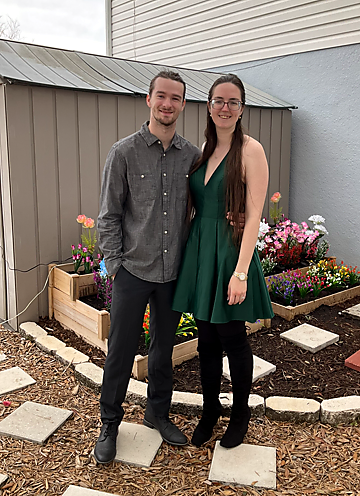 Man in a gray button-down shirt and black pants stands beside a woman in a green dress; they smile in a small garden with colorful flowers and a shed behind them.