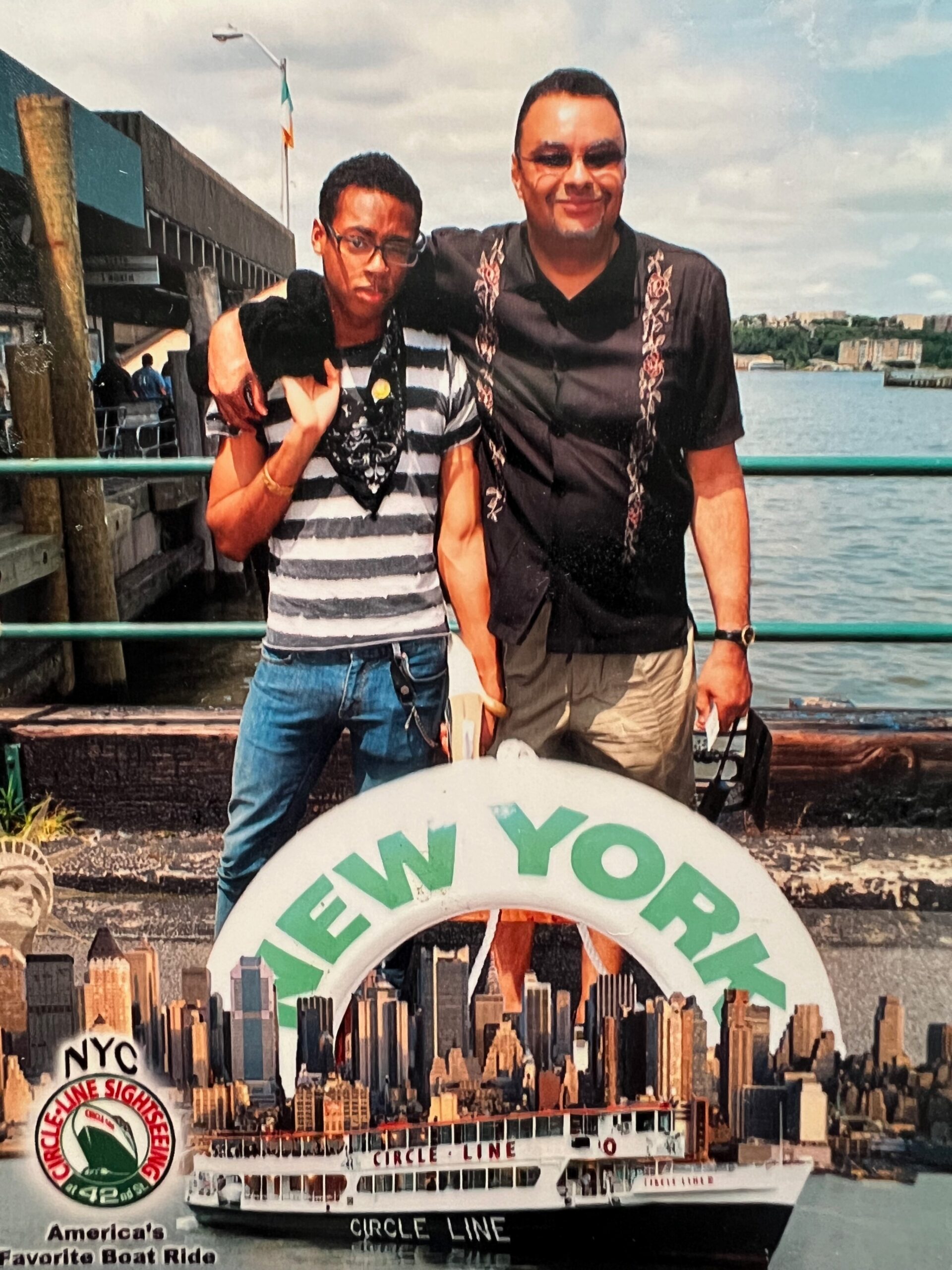 Two men posing together by the water with a Circle Line ferry and New York City skyline in the background.