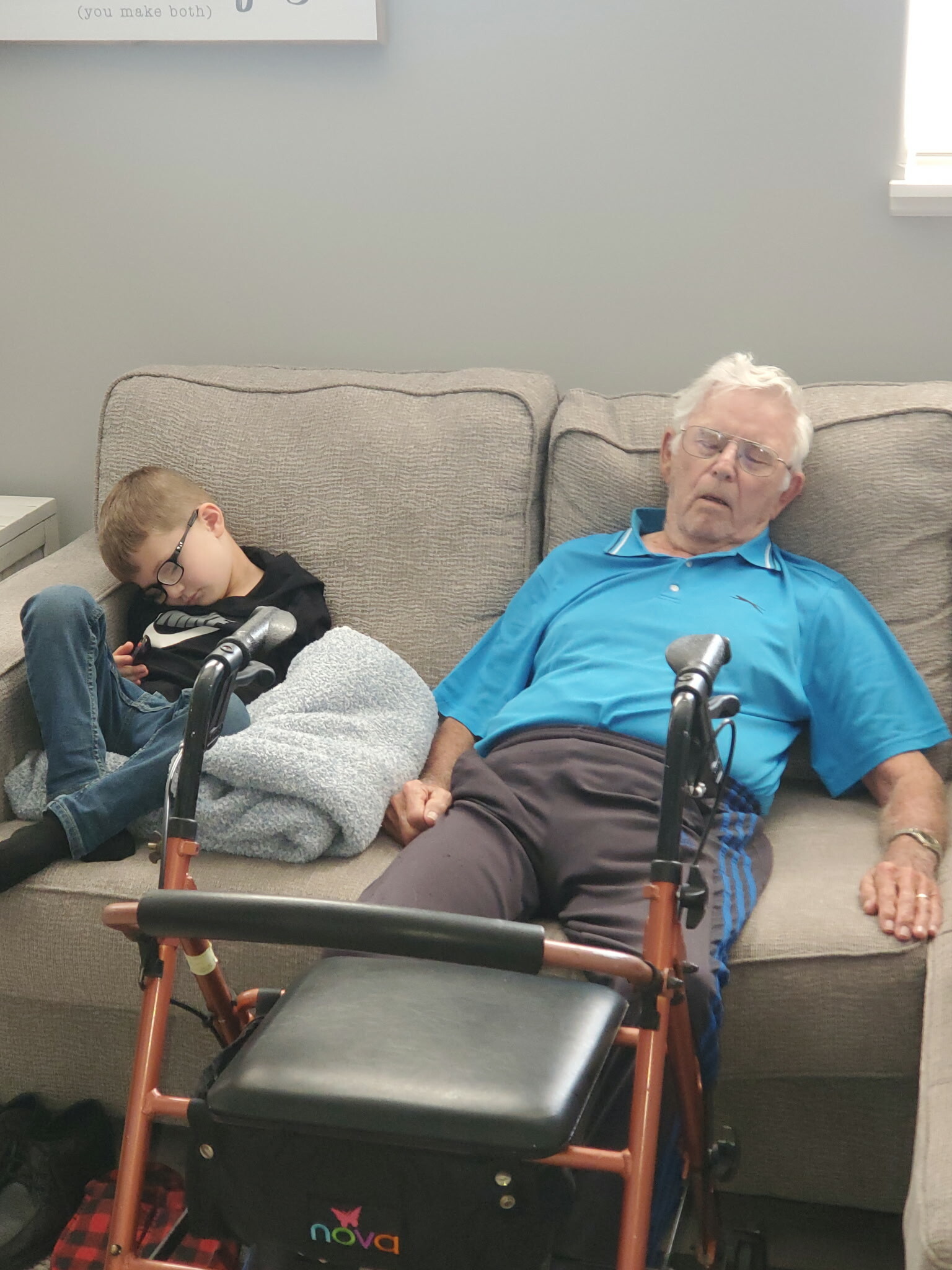 Elderly man in a blue polo shirt dozing on a gray couch beside a sleeping boy with glasses; a wooden-frame walker and a black bag sit in front.