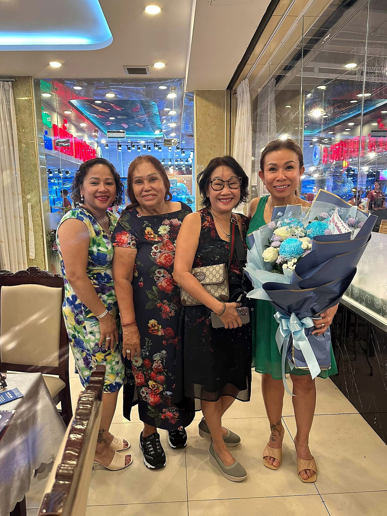 Four women posing together in a shop, one holding a large bouquet of blue and white flowers; bright floral dresses and casual sandals inside a glass-walled store.