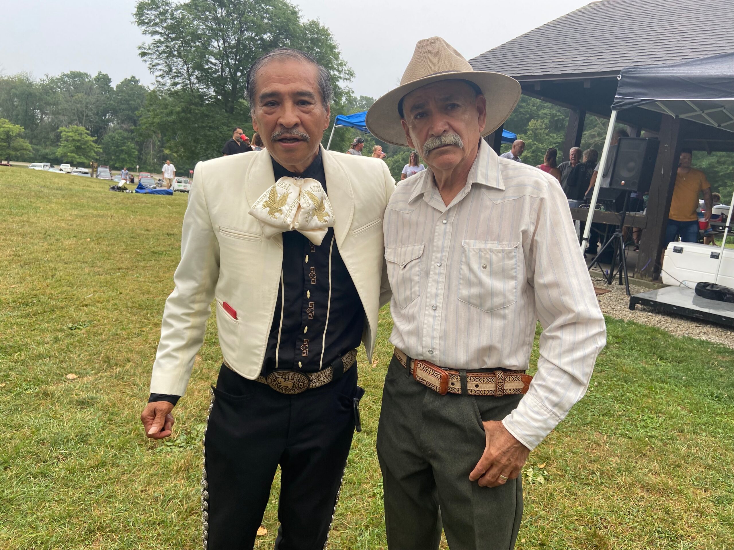 Two older men stand side by side at an outdoor event, wearing western-style outfits and belt buckles, looking at the camera.
