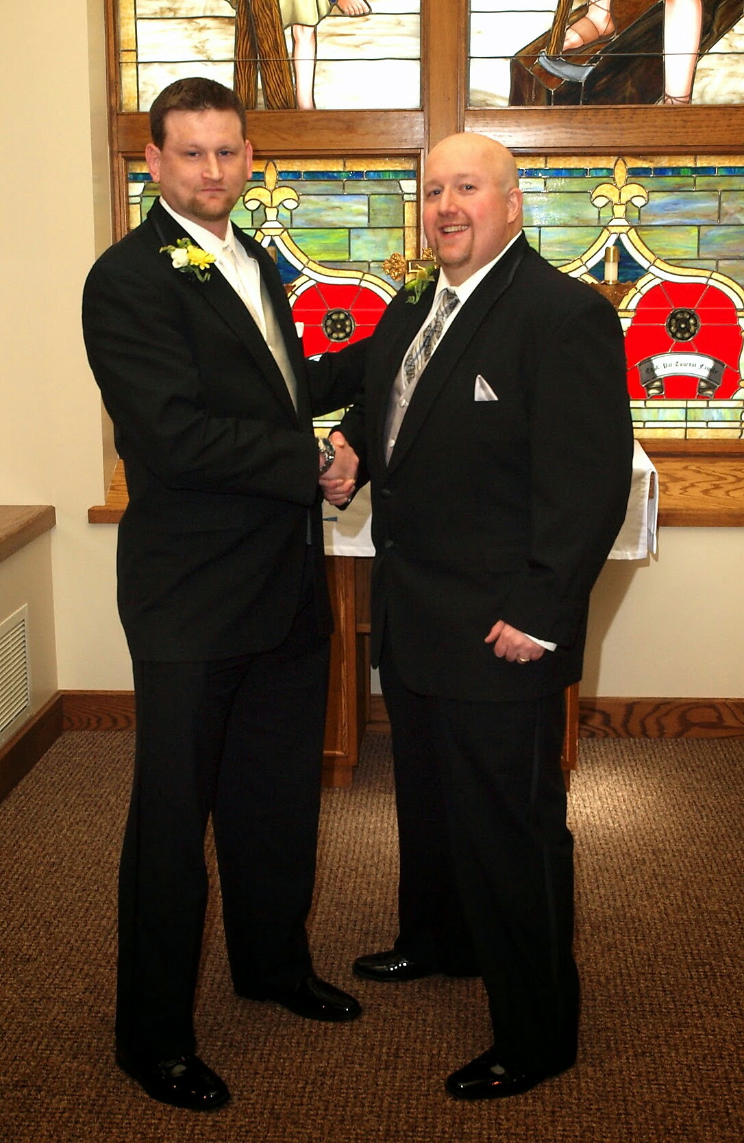 Two men in dark suits shake hands in front of a stained-glass window at a formal event in a church-like setting.
