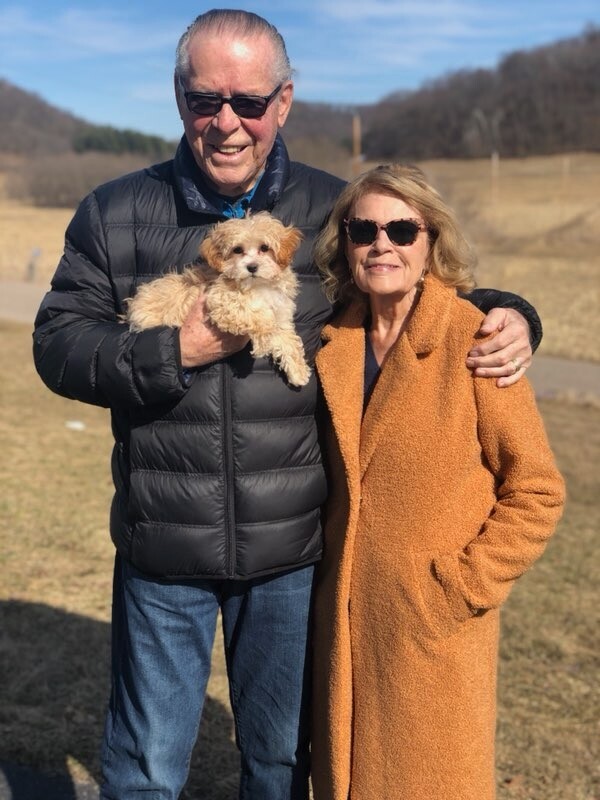 Older man in a black puffer jacket holds a small light-colored puppy; woman in an orange coat stands beside them outdoors, both wearing sunglasses under a sunny sky.