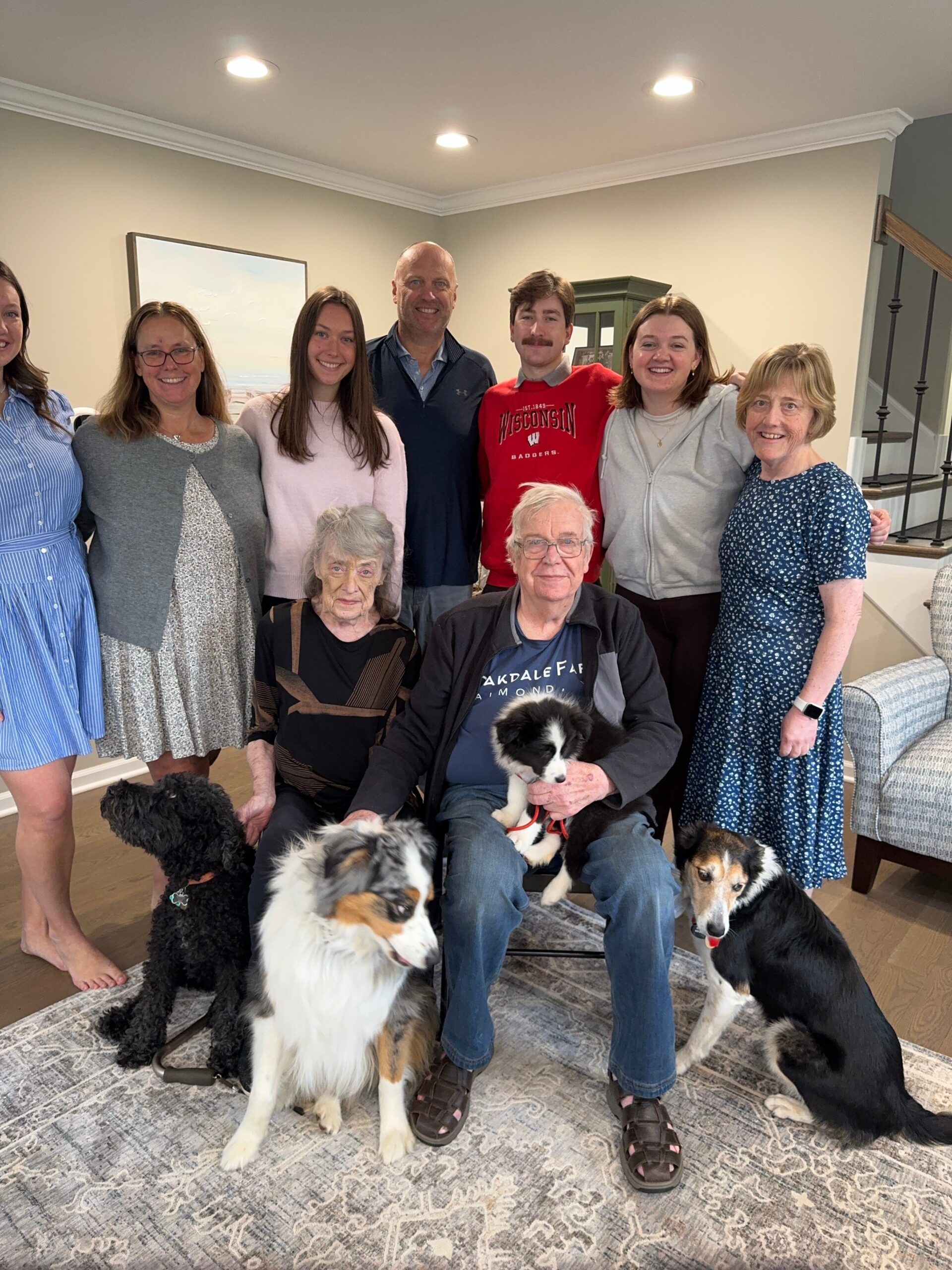 A group of nine adults posing together in a living room, smiling, with three dogs at their feet in front.
