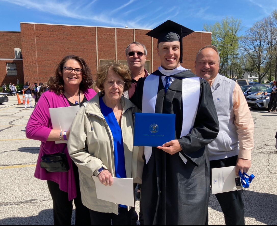 Graduate in cap and gown holding a blue diploma surrounded by family outdoors on graduation day in a parking lot. l