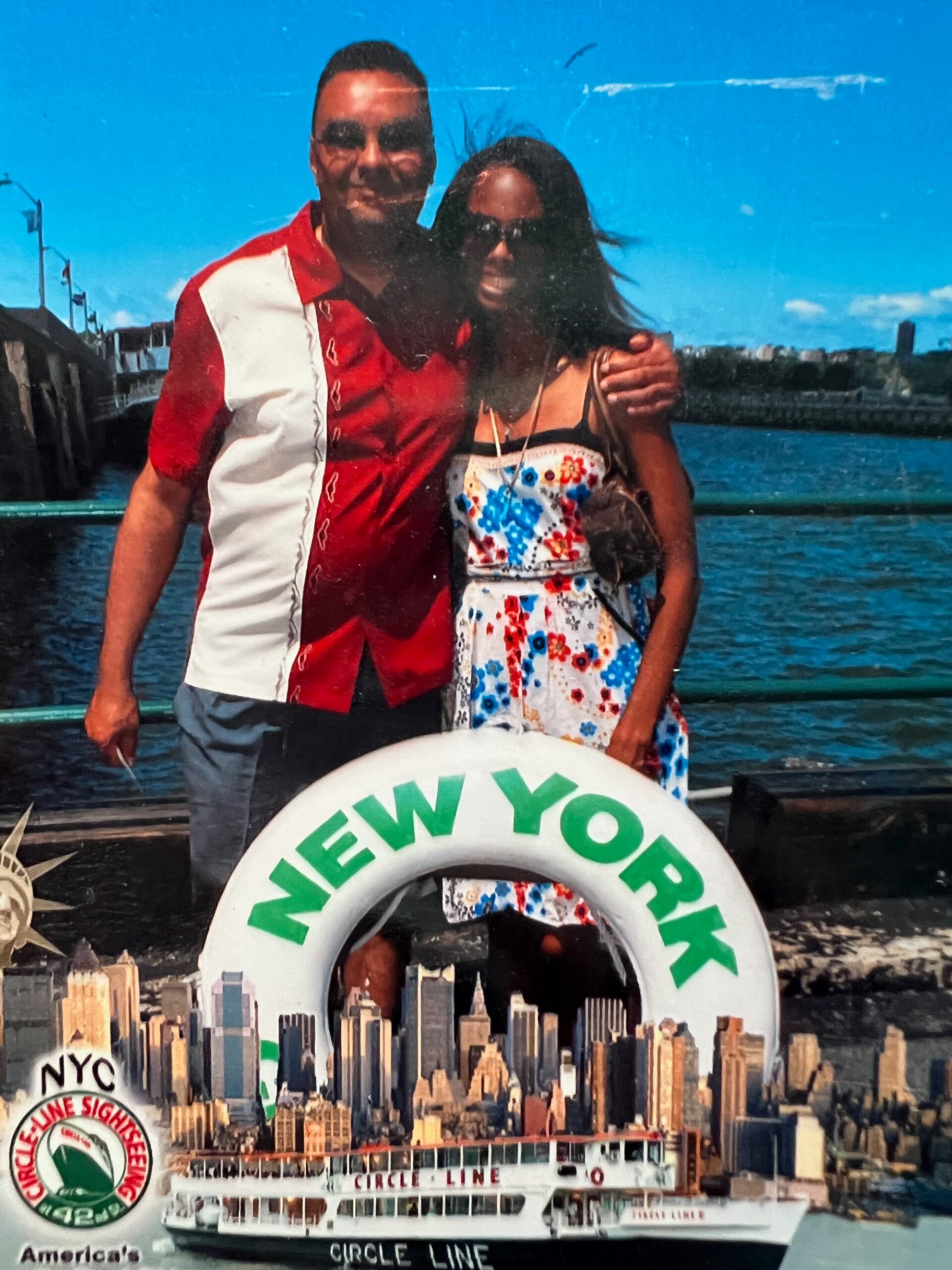 Couple posing together on a waterfront with a large New York circle line lifebuoy in the foreground and the NYC skyline in the background.
