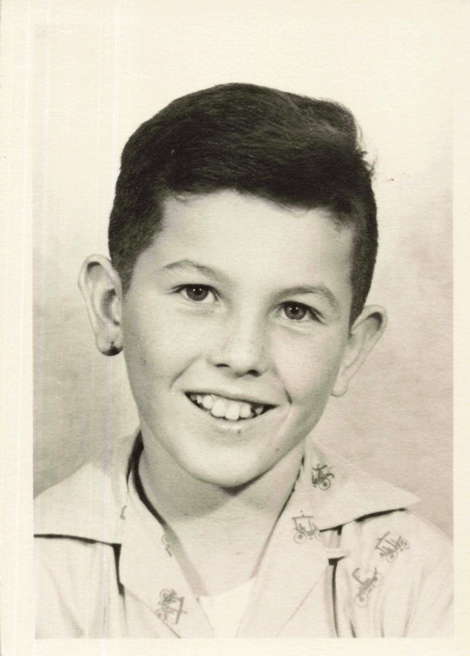 Black-and-white portrait of a smiling boy facing the camera, head and shoulders visible, wearing a light shirt with small print patterns.