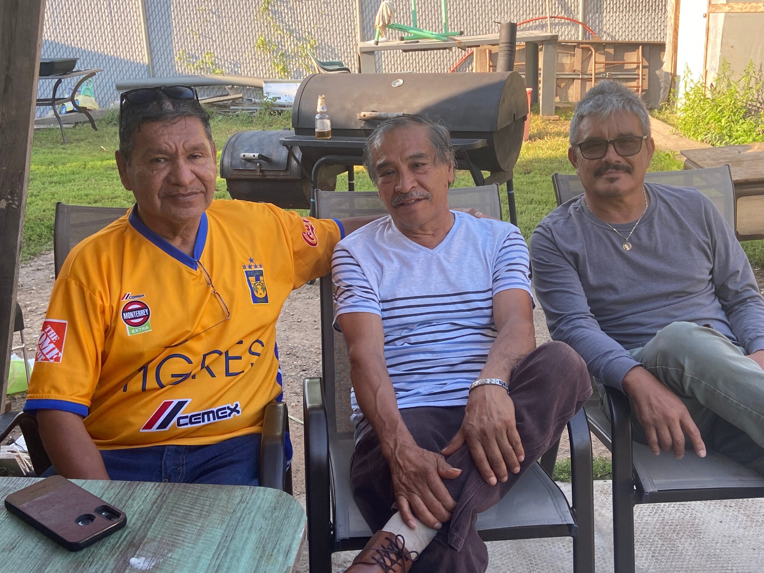 Three men sit together outdoors on patio chairs, one in a yellow sports jersey, one in a striped shirt, and one in a gray long‑sleeve shirt with glasses on top of his head, a grill behind them.