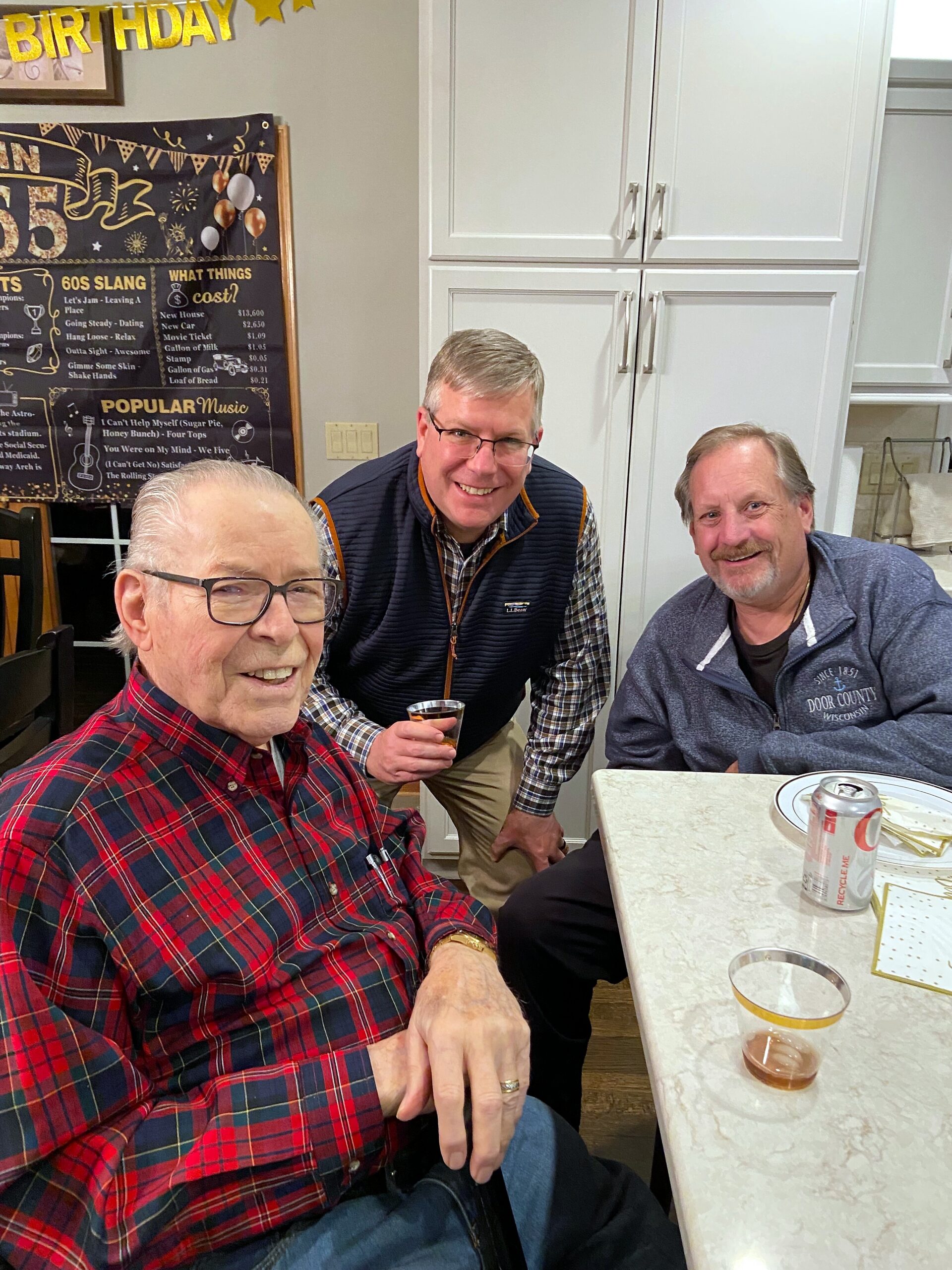 Three older men at a birthday gathering in a bright kitchen, smiling at the camera with drinks on the table in front of them.