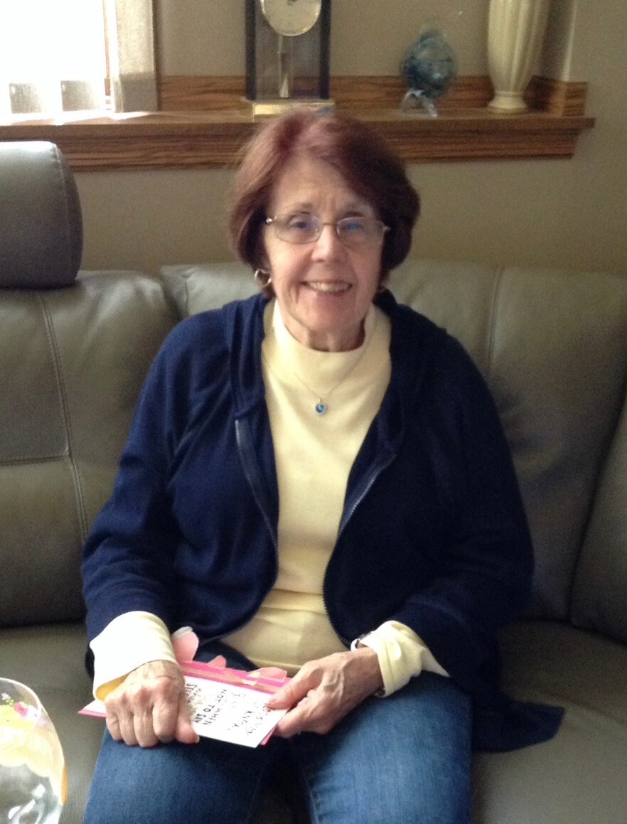 Smiling older woman with glasses sits on a gray couch, wearing a cream turtleneck and navy jacket, holding a pink handwritten card.