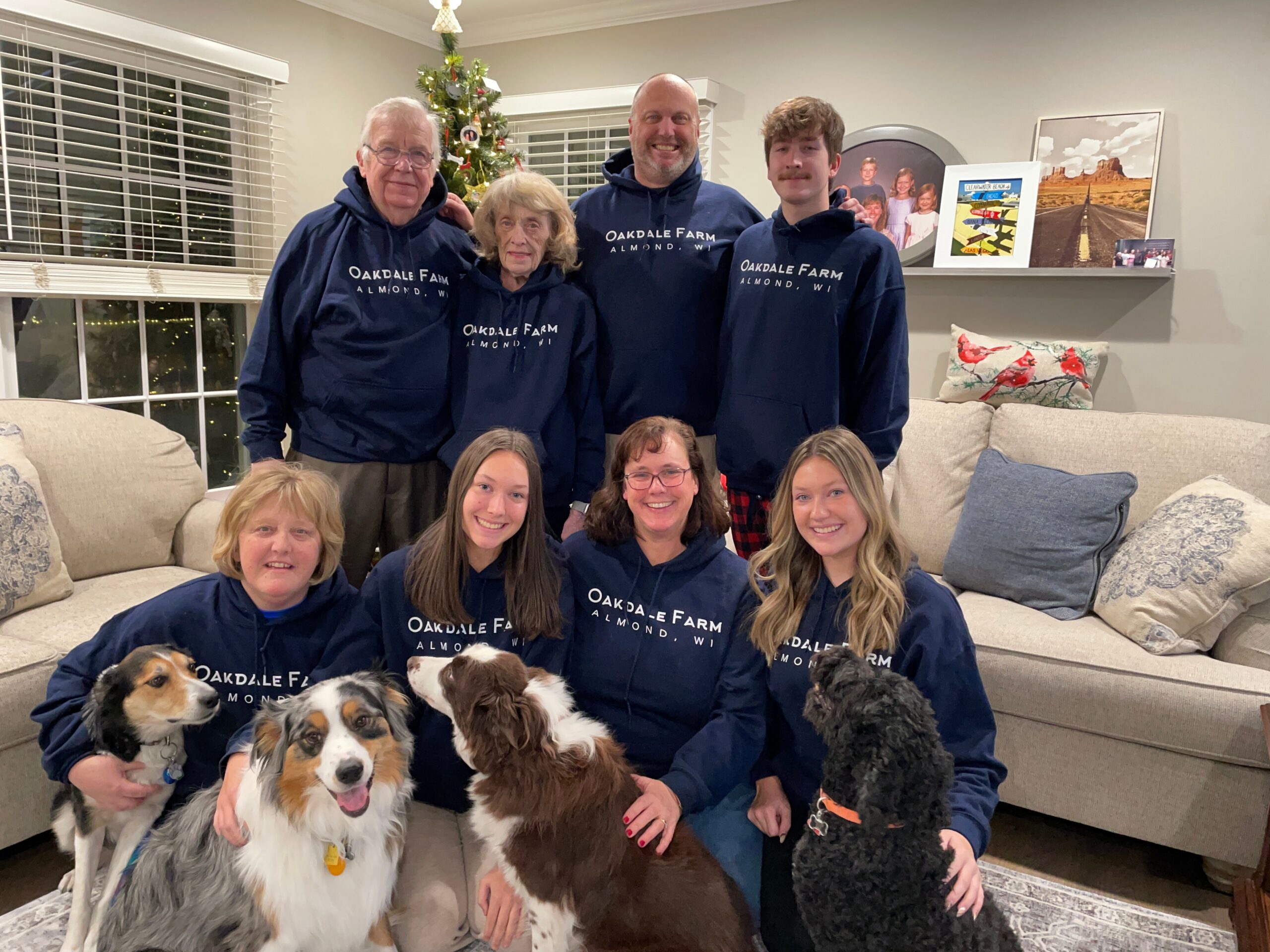 Family group photo in a living room with several dogs at their feet; everyone wears navy Oakdale Farm hoodies and smiles at the camera, Christmas tree in the background.