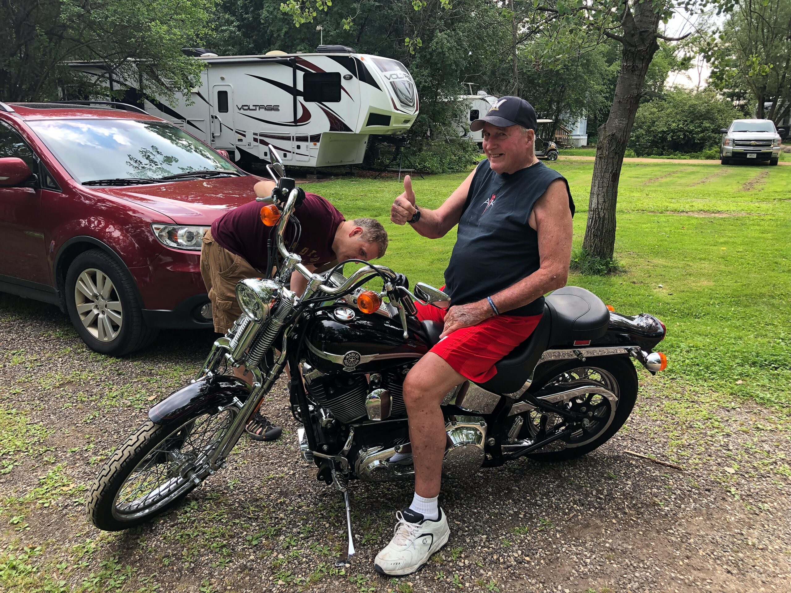 Two men with a motorcycle in a yard, one giving a thumbs up while an RV and a red SUV sit in the background.