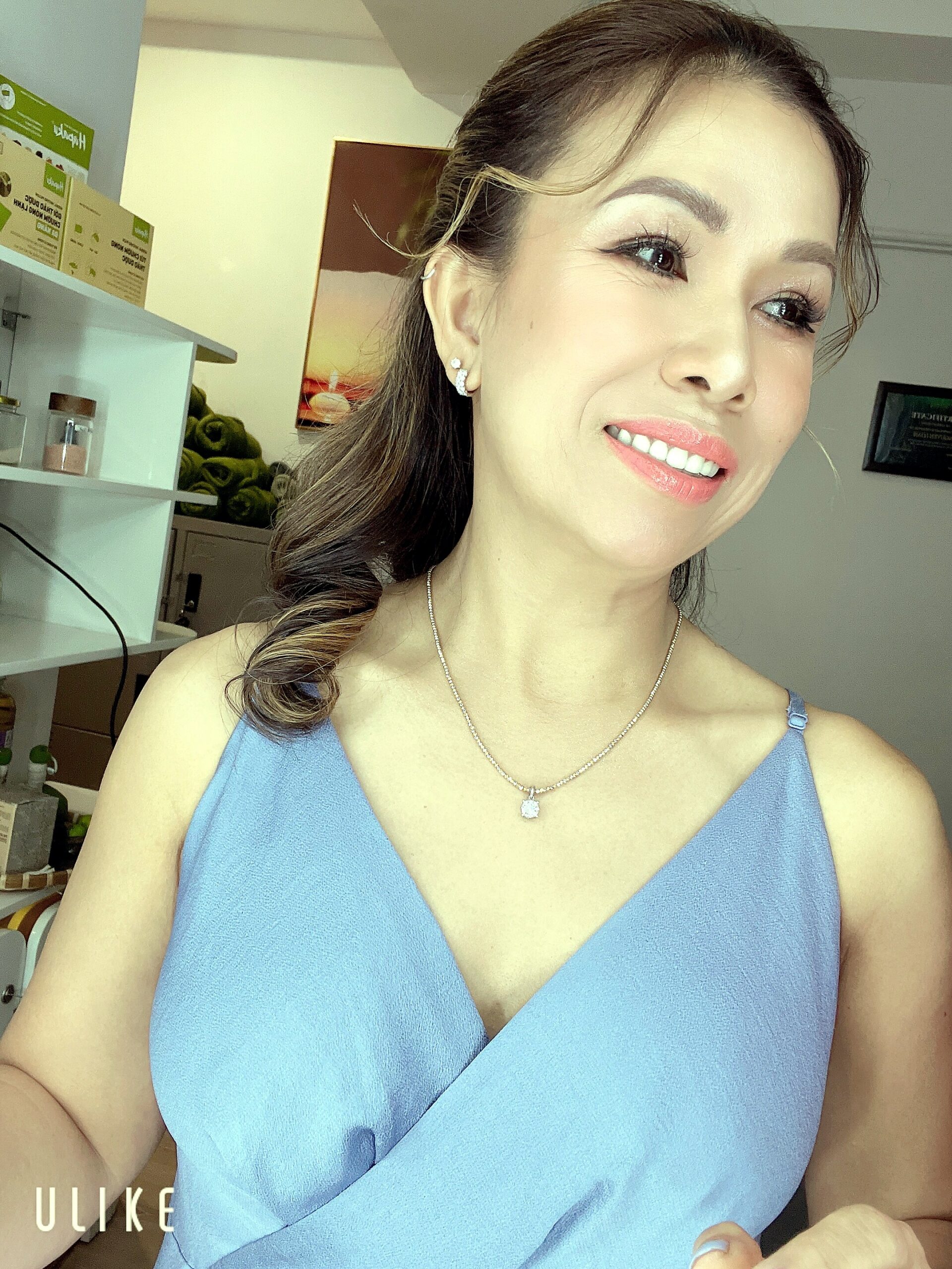 Close-up of a smiling woman in a light blue sleeveless top, wearing earrings and a pendant necklace in an indoor setting.