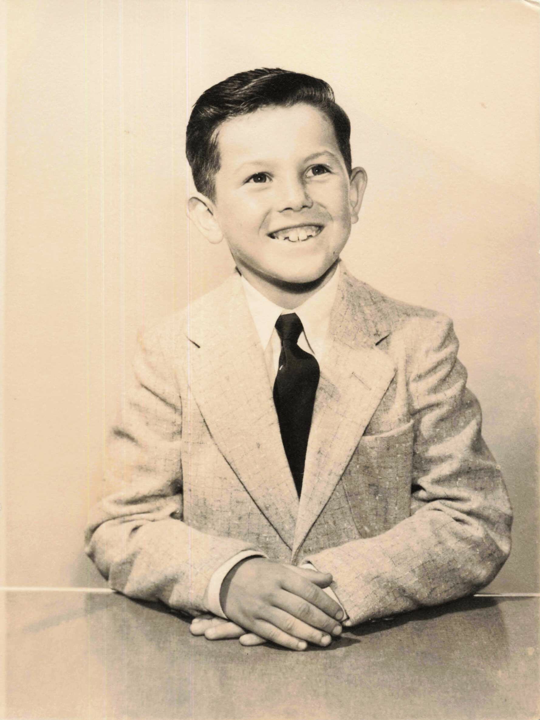 Young boy in a light suit and dark tie, smiling with hands folded on a table surface.