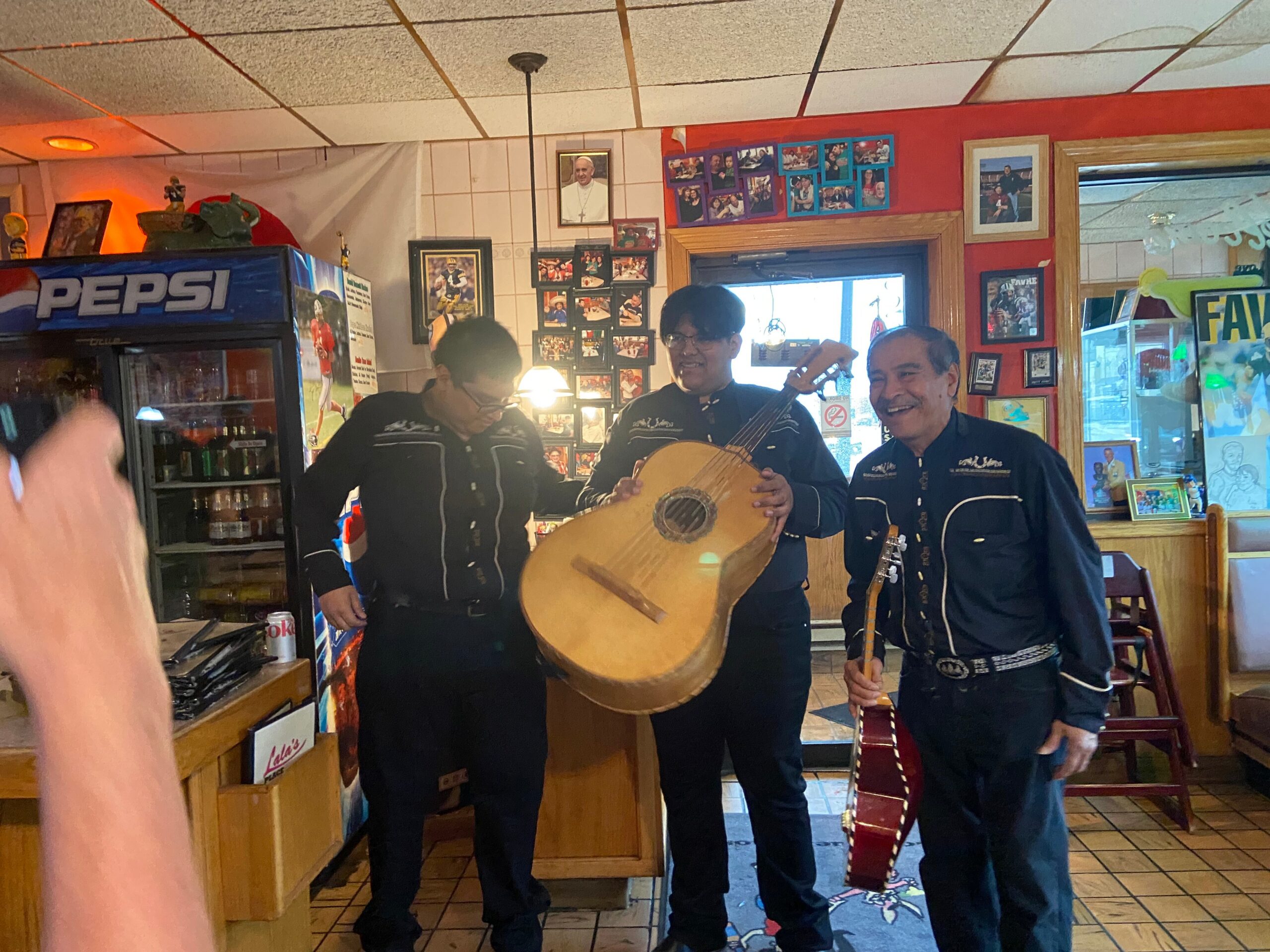 Three men in matching black jackets perform live music indoors, one holds an oud and another a guitar as a third smiles nearby.