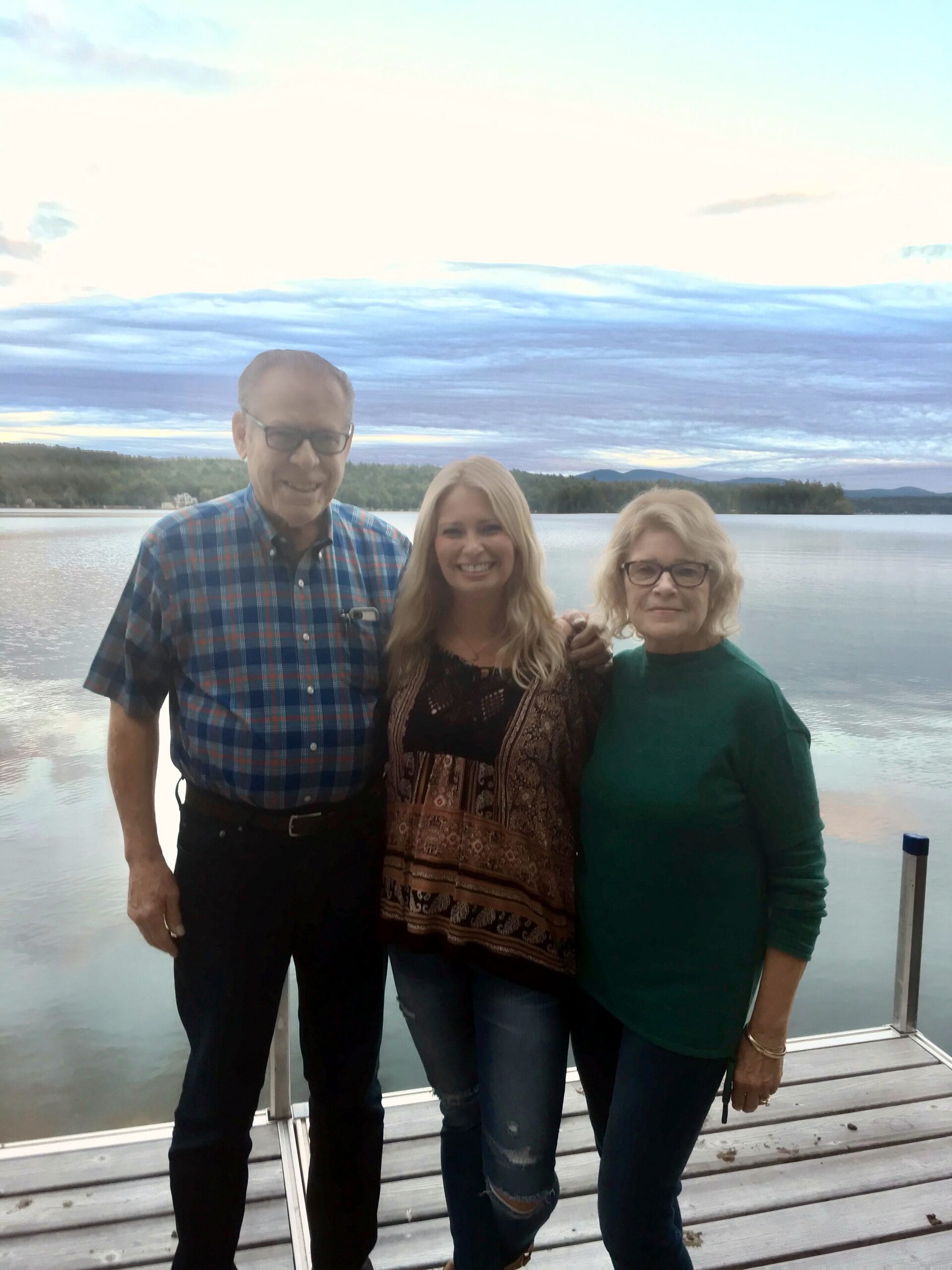Three adults stand on a wooden dock by a calm lake, smiling at the camera with a tree-lined shore in the background.