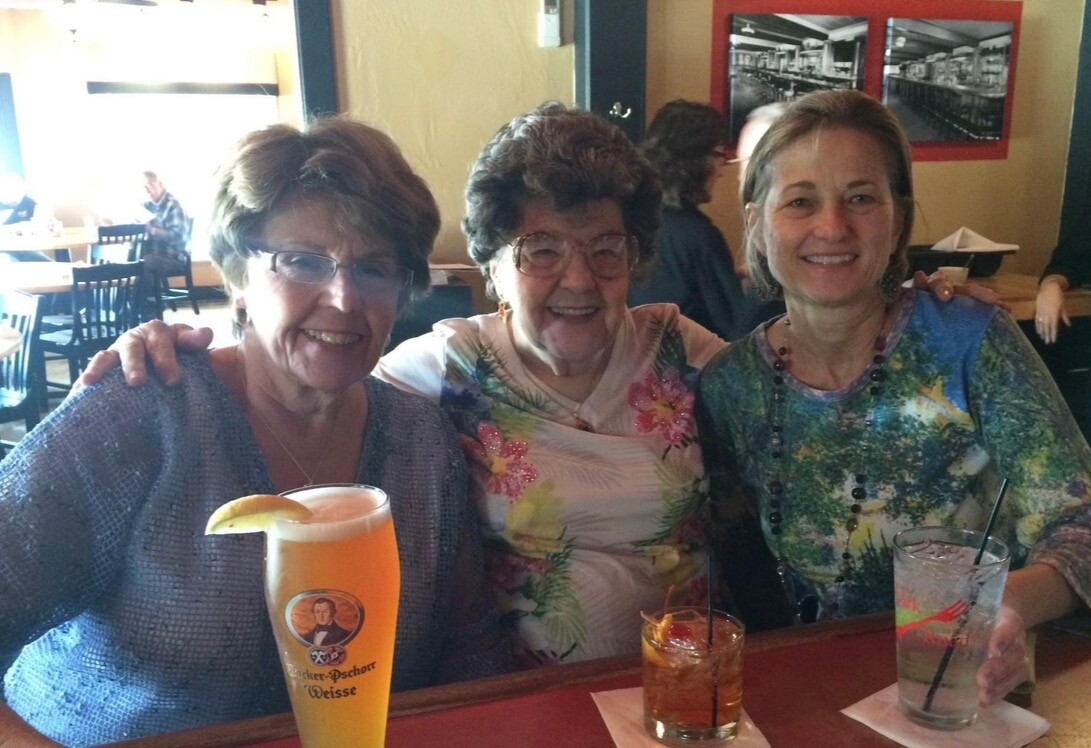Three women smile while seated at a restaurant bar, each with a different colorful drink in front of them.