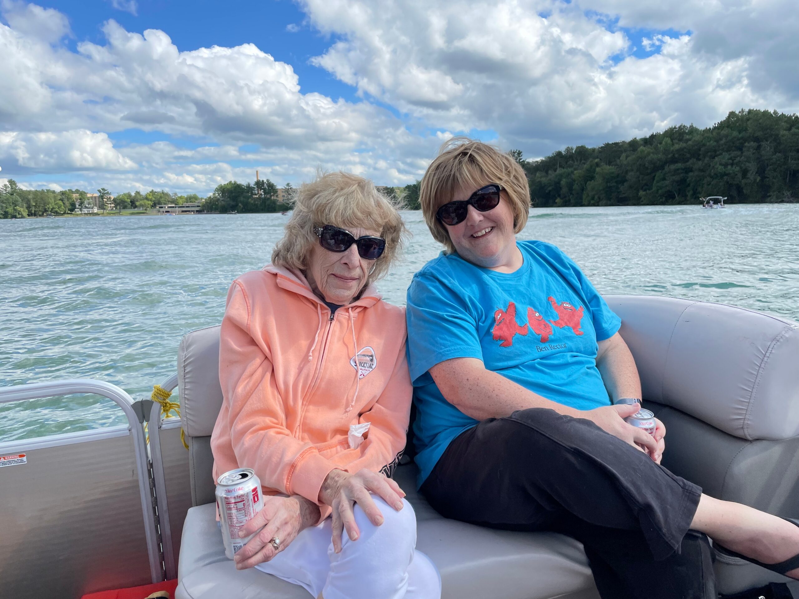 Two women wearing sunglasses sit together on a boat on a lake, smiling with a blue sky and clouds behind them.